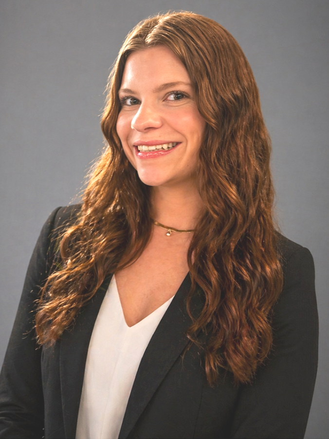 Portrait of a young woman with long, wavy, auburn hair, smiling, wearing a black blazer over a white top, and a delicate choker necklace, against a plain gray background.