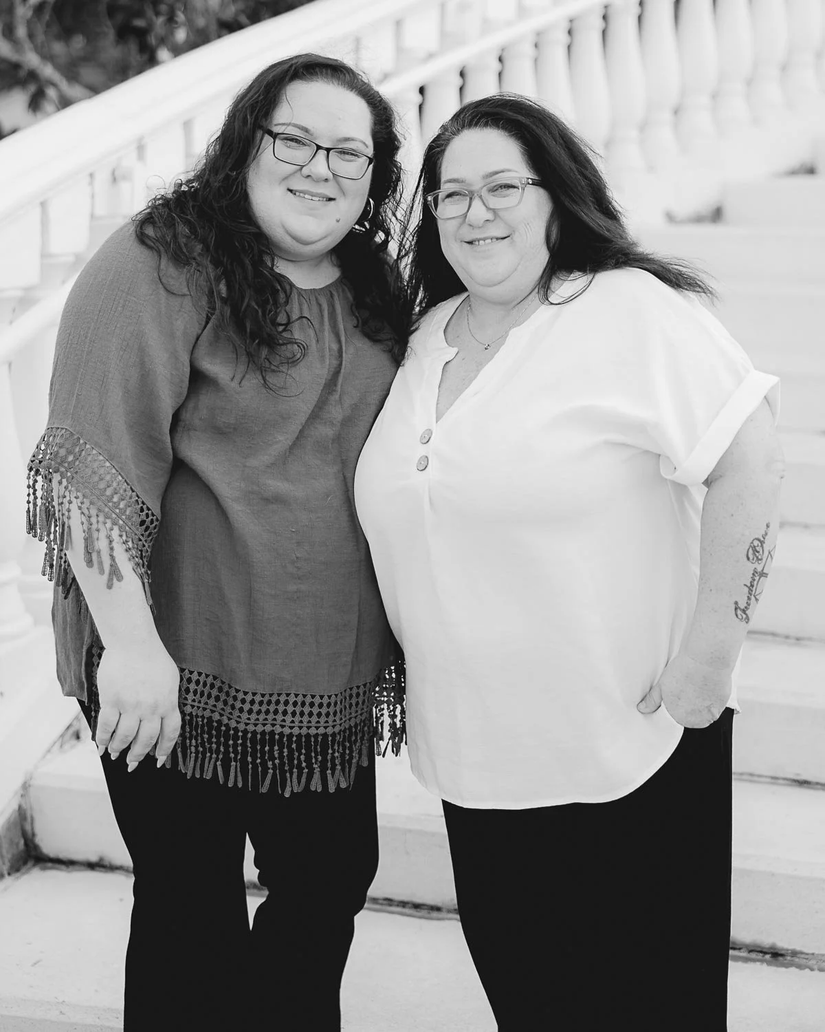 Two women smiling and standing close together on a staircase with a railing in the background, in black and white.