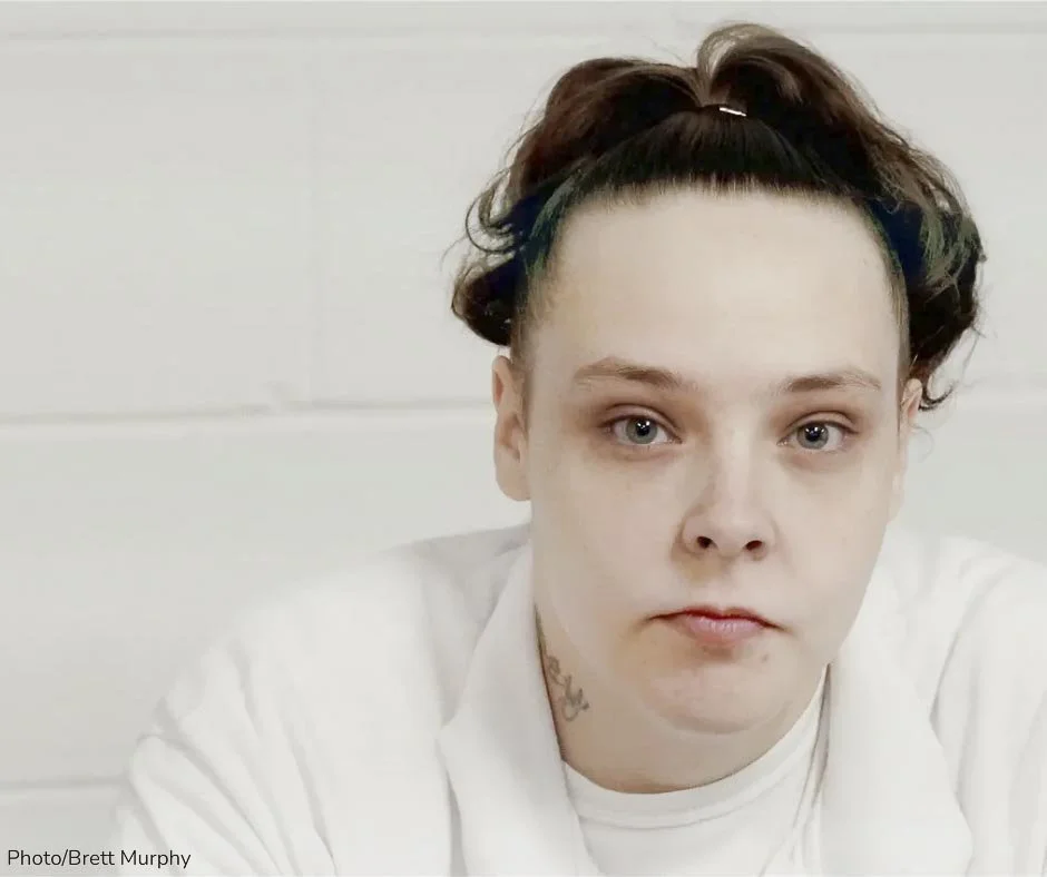 Close-up of a young woman with a serious expression, wearing a white shirt, and sporting a brown curly hairdo with a headband. She has gray eyes, a tattoo on her neck, and is sitting against a white background.