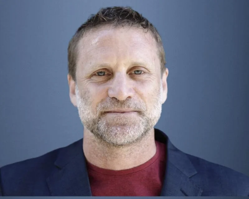Close-up of a middle-aged man with short brown hair, blue eyes, and a beard, wearing a dark blazer over a maroon shirt against a solid dark background.