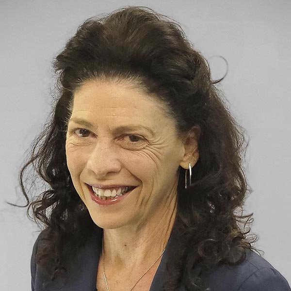 A woman with dark curly hair, wearing a dark blazer, silver hoop earrings, and a necklace, smiling at the camera against a neutral gray background.