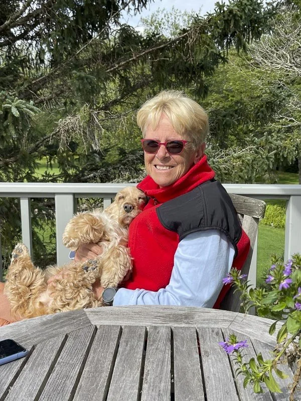 An older woman with blonde hair, wearing pink sunglasses and a red vest, sitting outside on a deck holding a small, curly-haired dog.