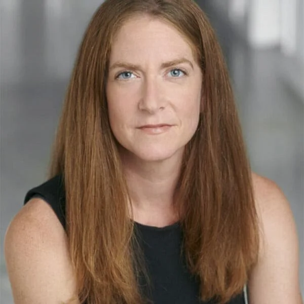A woman with long red hair, blue eyes, and fair skin, wearing a black sleeveless top, sitting in an indoor setting with a blurred background.