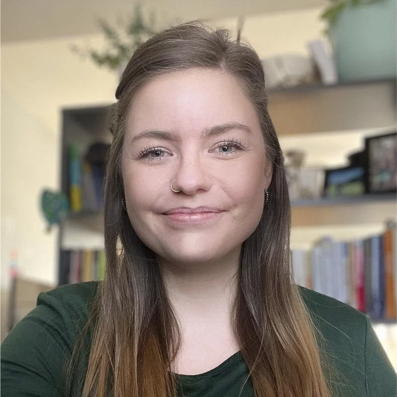 Young woman with long brown hair, wearing a green top, smiling, in a room with bookshelves and plants in the background.