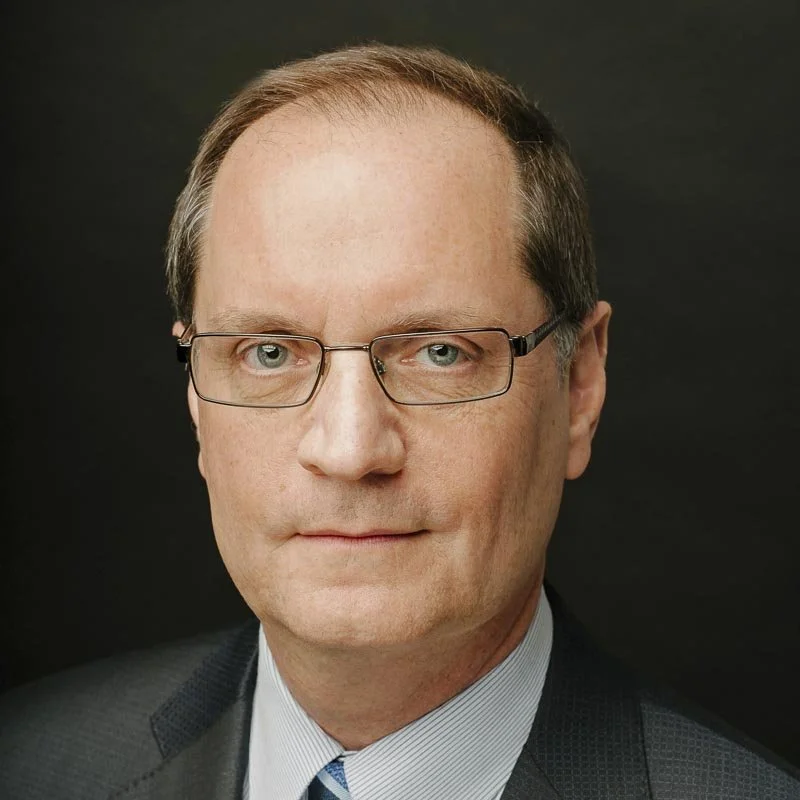 A middle-aged man wearing glasses, a dark suit, and a light blue striped shirt against a black background.