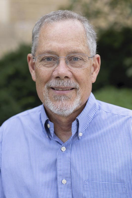A smiling middle-aged man with glasses and a beard in a blue checkered shirt standing outdoors with greenery in the background.