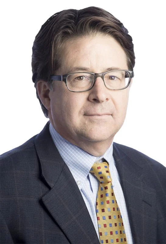 Portrait of a middle-aged man with dark hair, glasses, wearing a business suit, light blue shirt, and patterned yellow tie, against a white background.