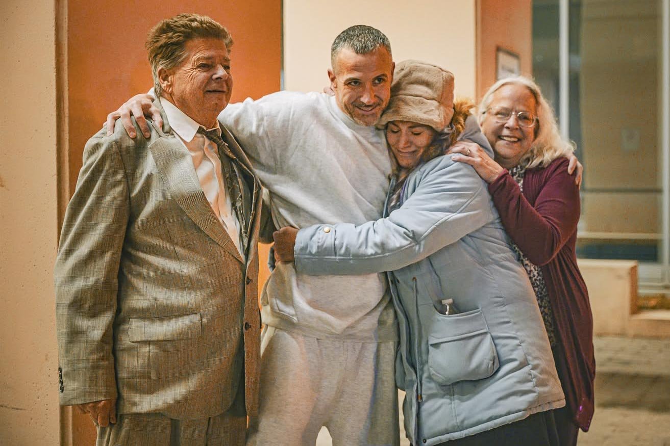 Four adults, two men and two women, embrace and smile together indoors, showing a moment of happiness and connection.