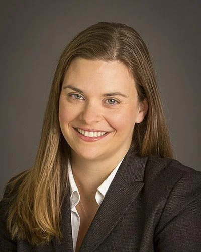 A woman with long brown hair wearing a black blazer and white shirt smiles at the camera against a gray background.