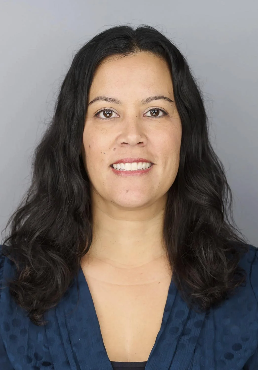A woman with long, wavy black hair, smiling, wearing a dark blue top, against a gray background.
