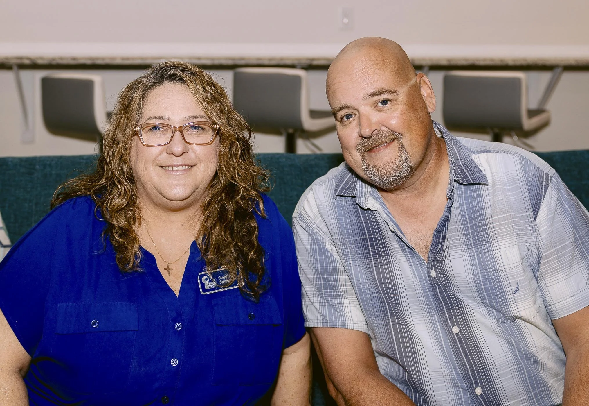 A woman with curly hair and glasses wearing a blue shirt and a man with a beard and bald head wearing a gray plaid shirt sitting together on a teal couch in an indoor setting.