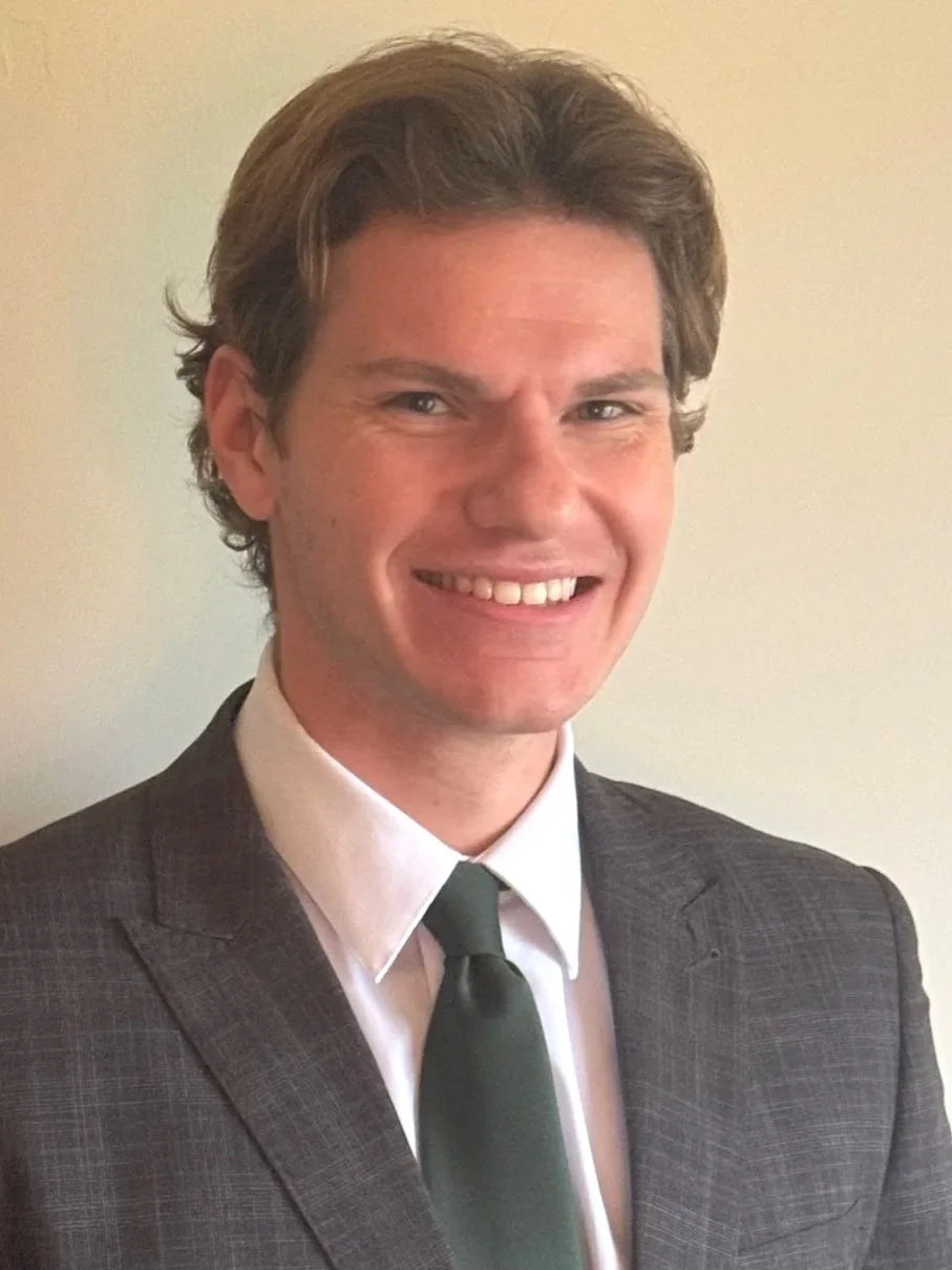 A young man with light skin, brown hair, wearing a dark suit, white shirt, and green tie, smiling and looking at the camera against a plain light-colored wall.