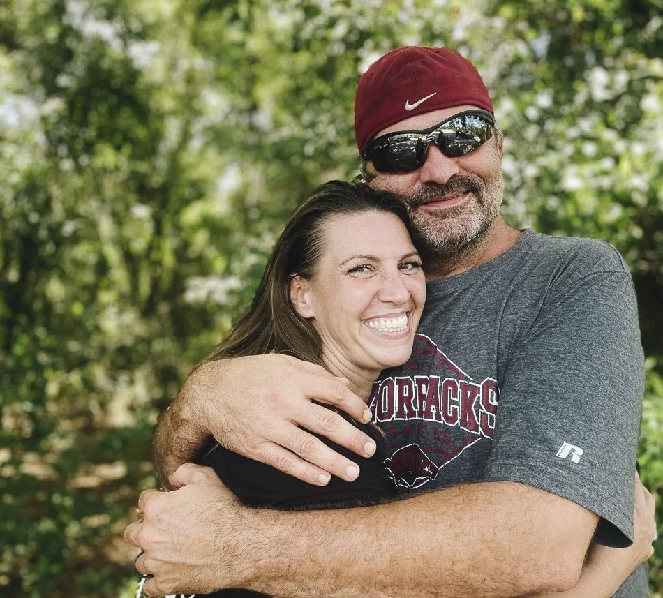 A man wearing a red baseball cap, sunglasses, and a gray t-shirt hugging a woman with long brown hair, smiling in a green outdoor setting.