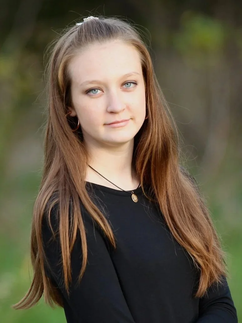 A young woman with long, light brown hair and blue eyes standing outdoors in front of a blurred green background. She is wearing a black top, a necklace with a small pendant, and hoop earrings.