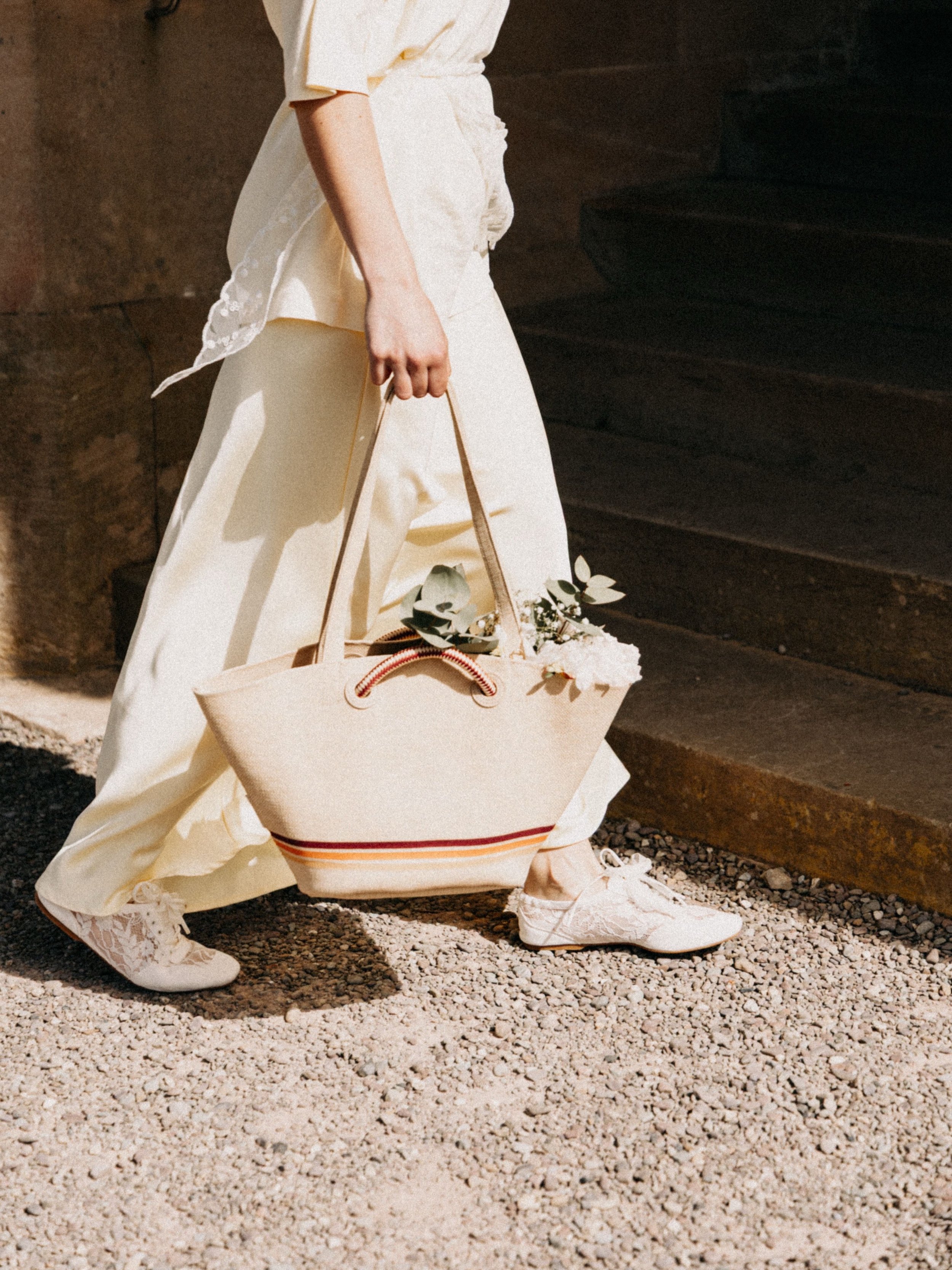 Person dressed in white clothing, holding a large beige bag with a striped pattern and some greenery, standing on gravel near dark steps.