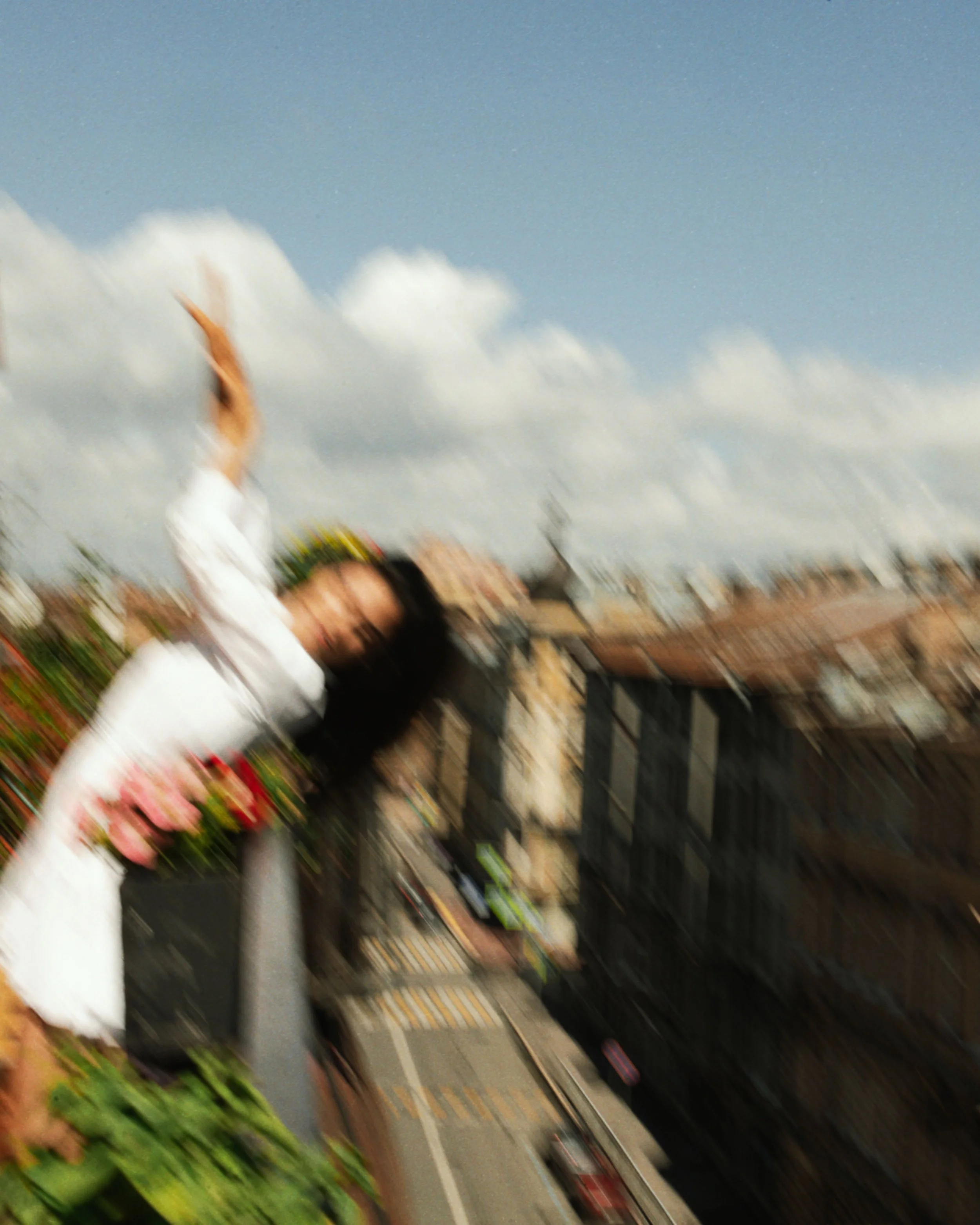 Blurred image of a woman wearing white clothing and a headscarf, standing in front of shelves stocked with various products, in what appears to be a grocery store aisle. The background shows a partly cloudy sky.