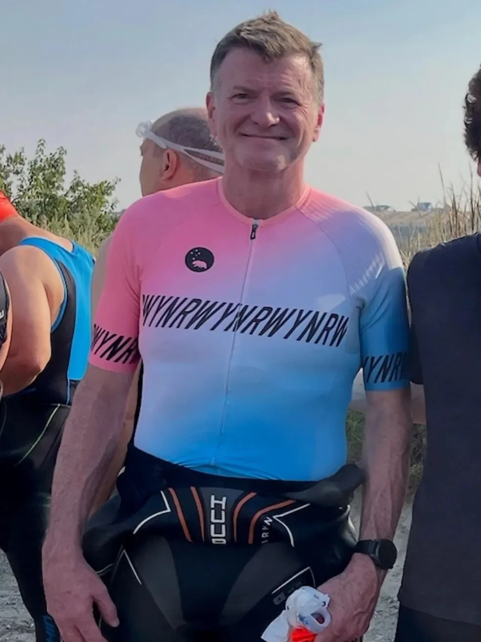 A smiling man wearing a pink, white, and light blue gradient cycling jersey with black text, standing outdoors with other cyclists around him, during daytime.
