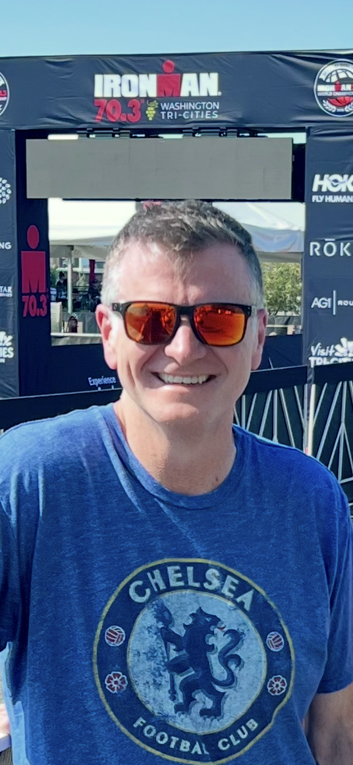 Smiling man wearing sunglasses and a Chelsea Football Club t-shirt at the IRONMAN 70.3 Washington Tri-Cities event.