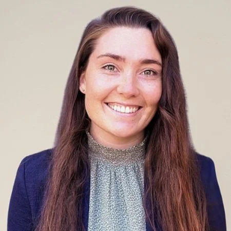 A young woman with long brown hair, smiling, wearing a blue blazer and a gray blouse, standing against a neutral background.