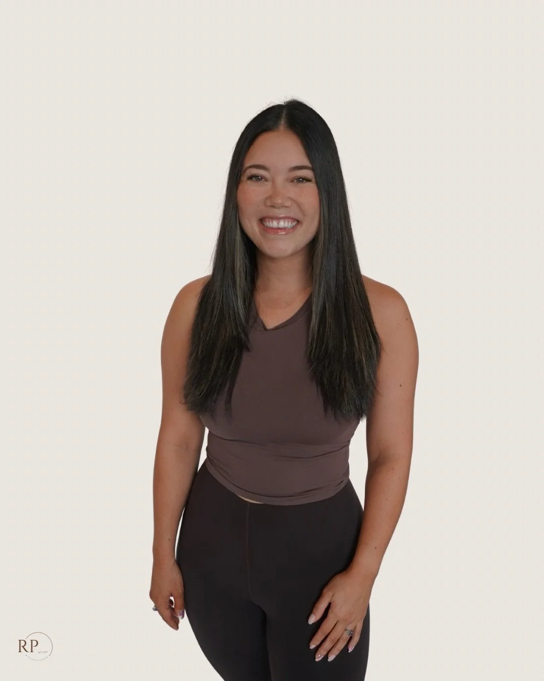 Woman with long dark hair smiling in a sleeveless brown top and black pants against a plain background.