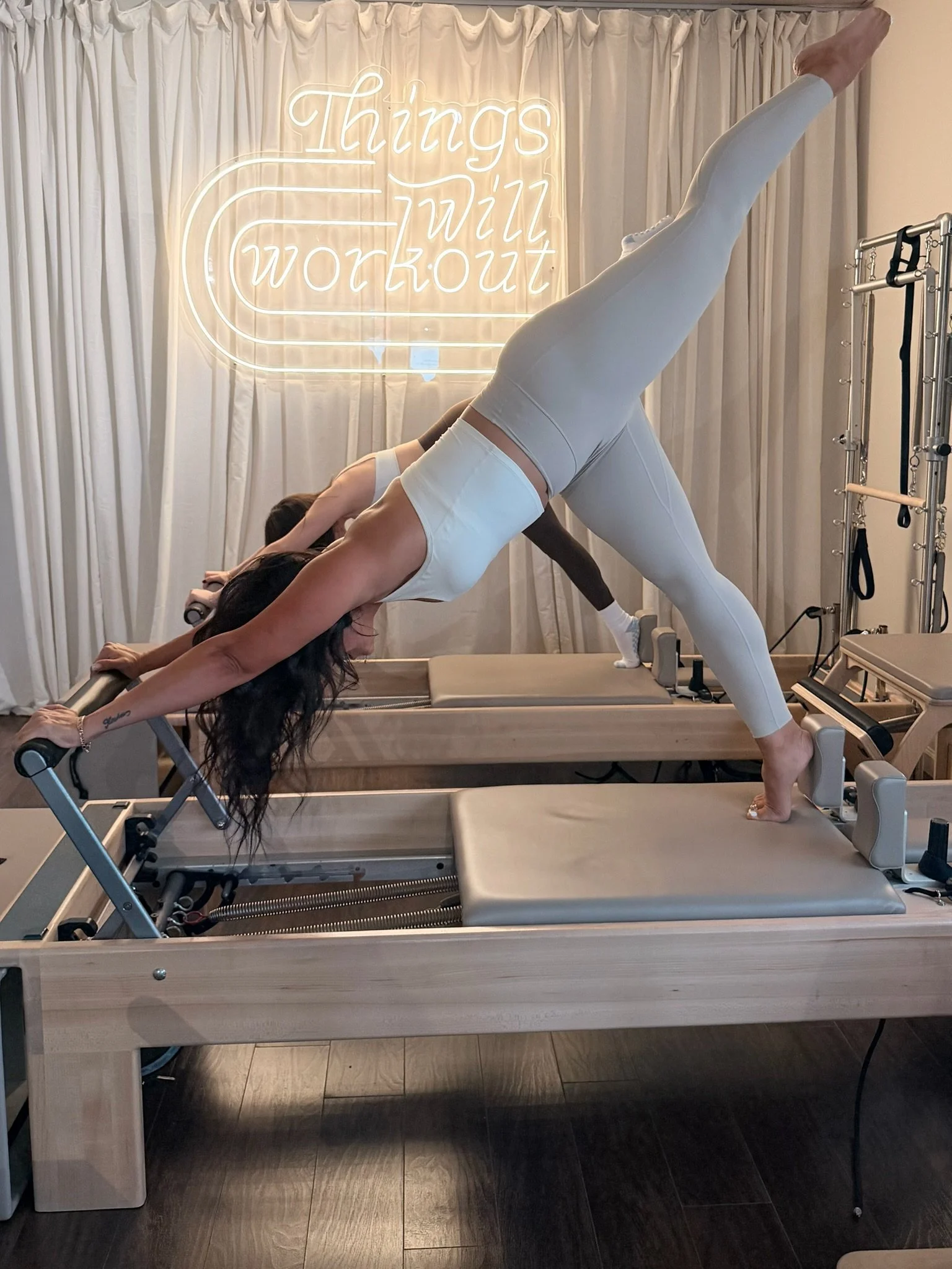 A woman doing a plank yoga pose on a Pilates reformer machine in a fitness studio with a cream-colored curtain and a neon sign that reads 'Things Will Work Out' in the background.