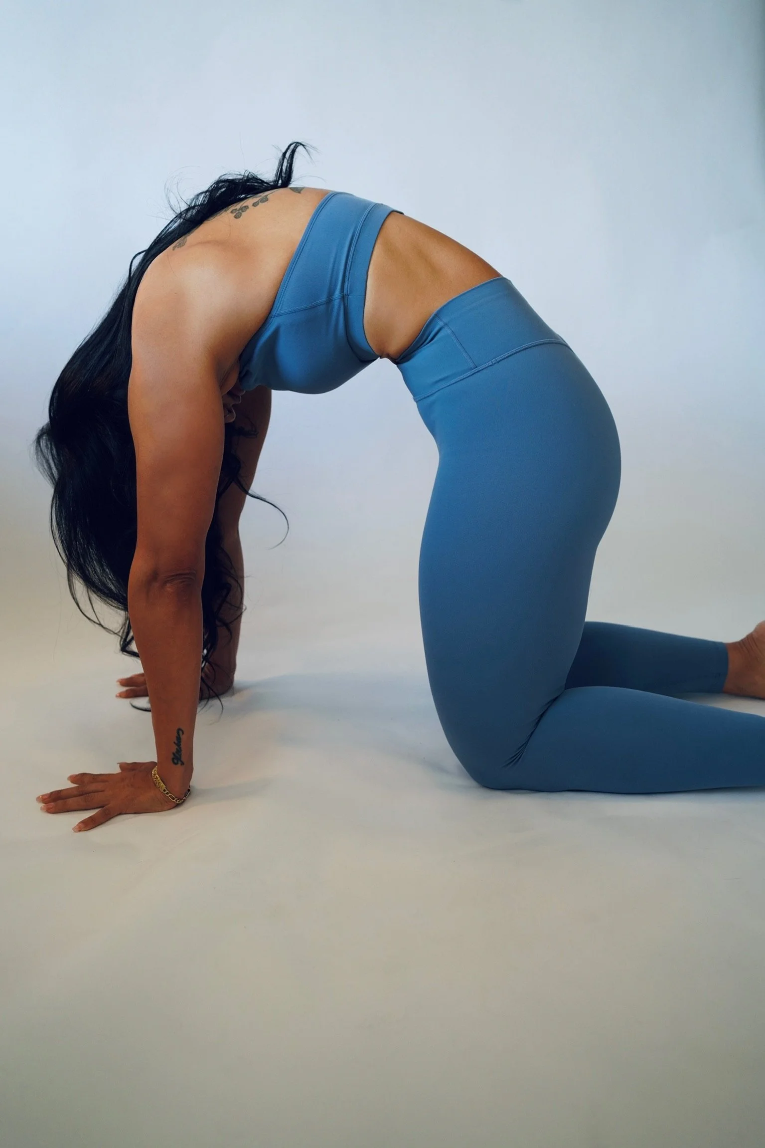 A woman practicing yoga, in a crouched position with hands and knees on the floor, wearing a blue sports bra and matching leggings, with long black hair and tattoos on her arm.