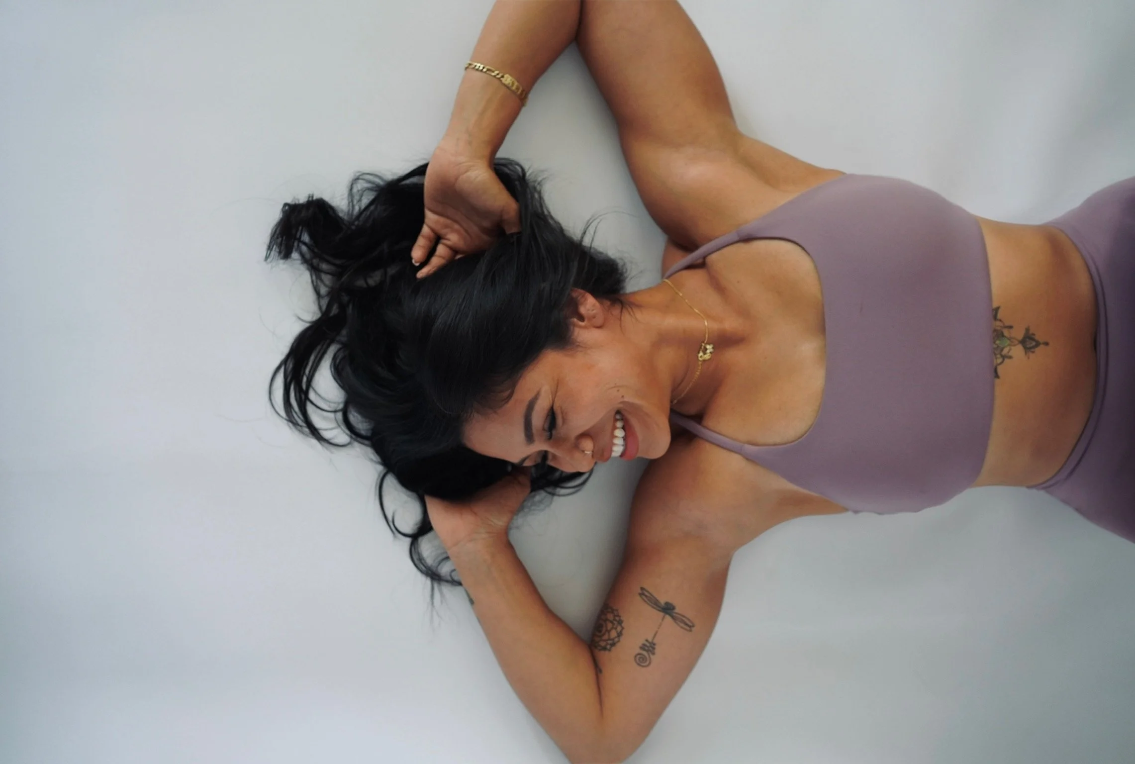 A woman with dark hair, tattoos, and jewelry, smiling while lying on the floor with her eyes closed, wearing a purple tank top and beige shorts.
