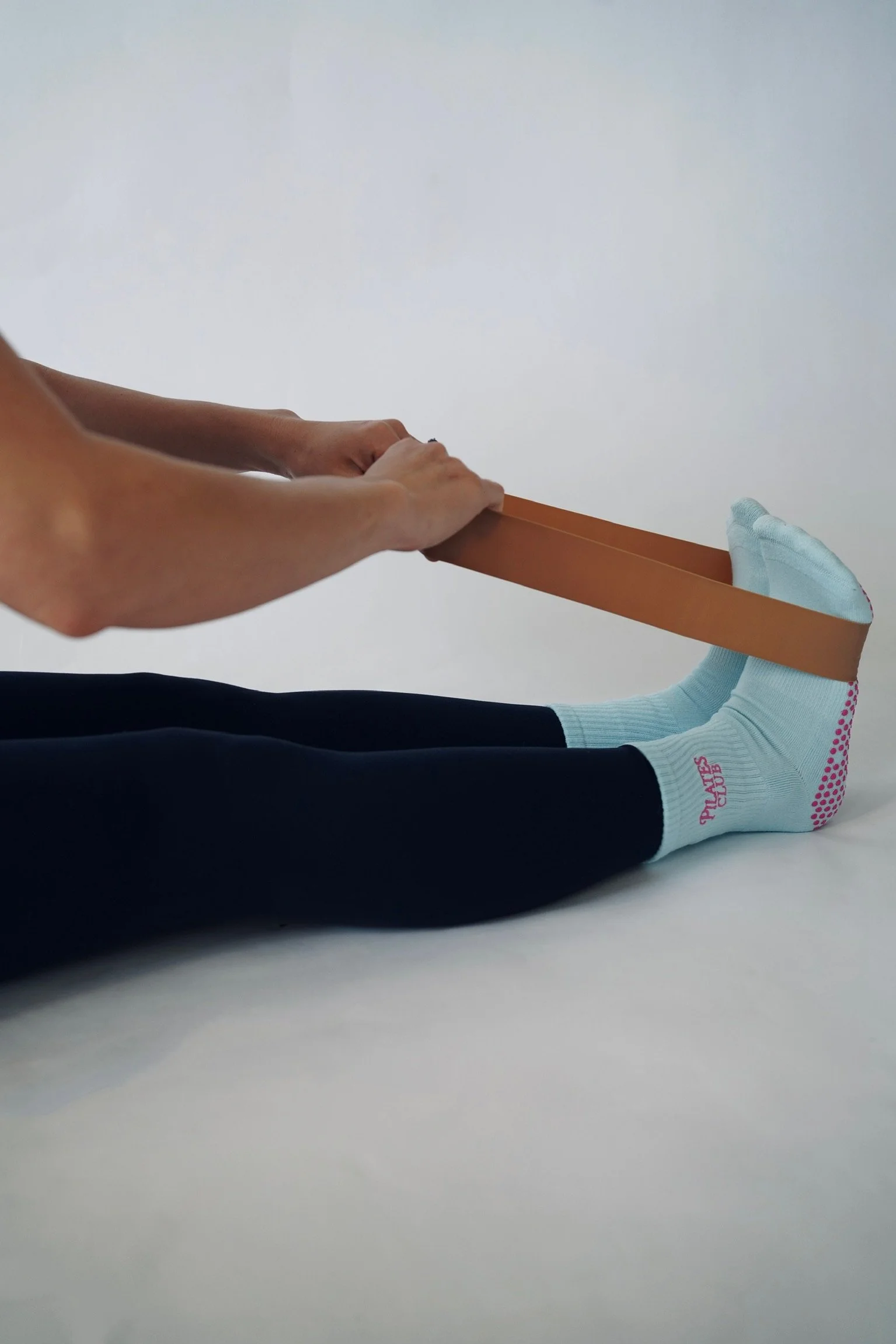 Person stretching with legs extended, holding a wooden stick, wearing black pants and light blue socks, lying on a white surface