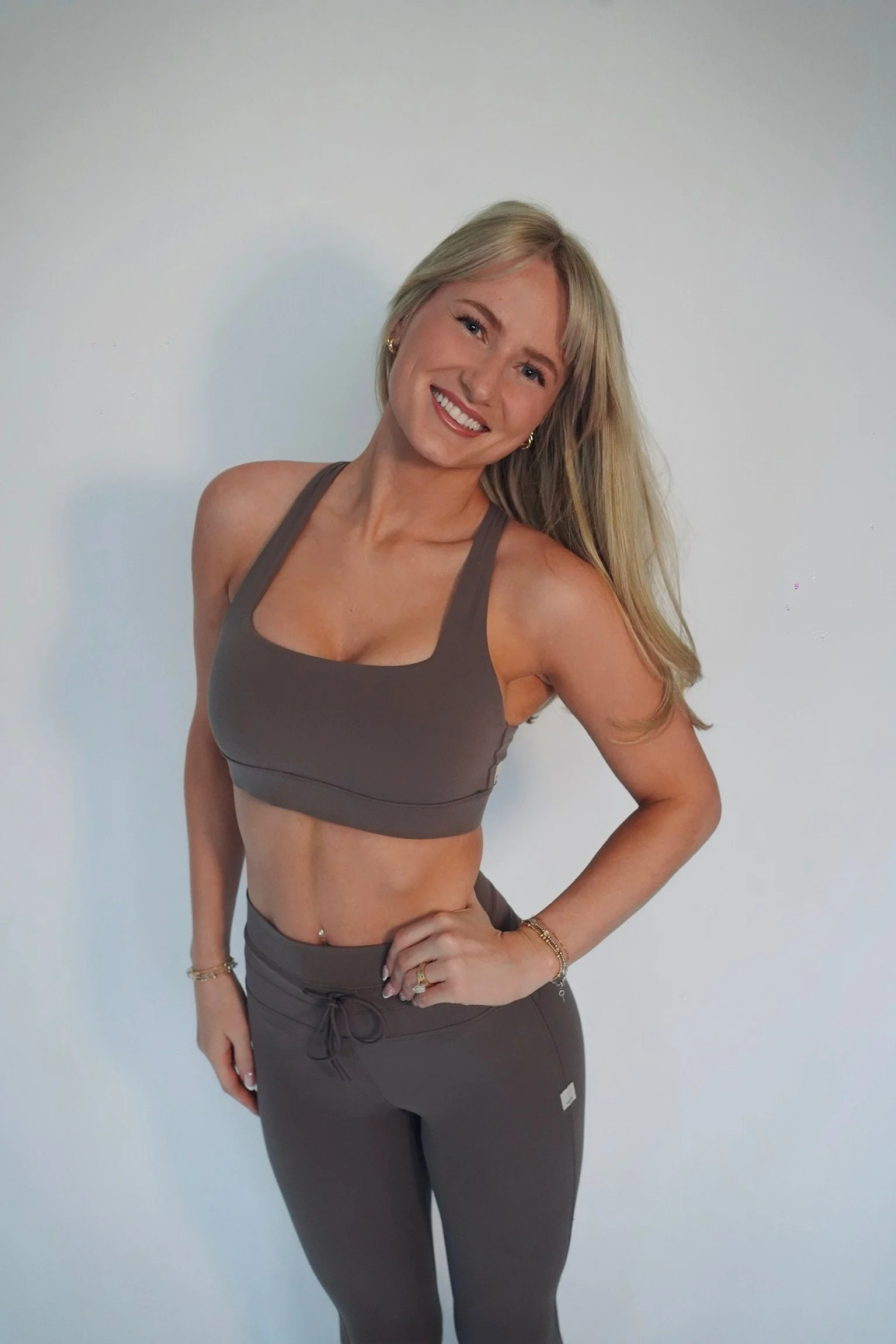 A smiling woman with long blond hair, wearing a gray sports bra and matching workout pants, standing against a plain white wall.