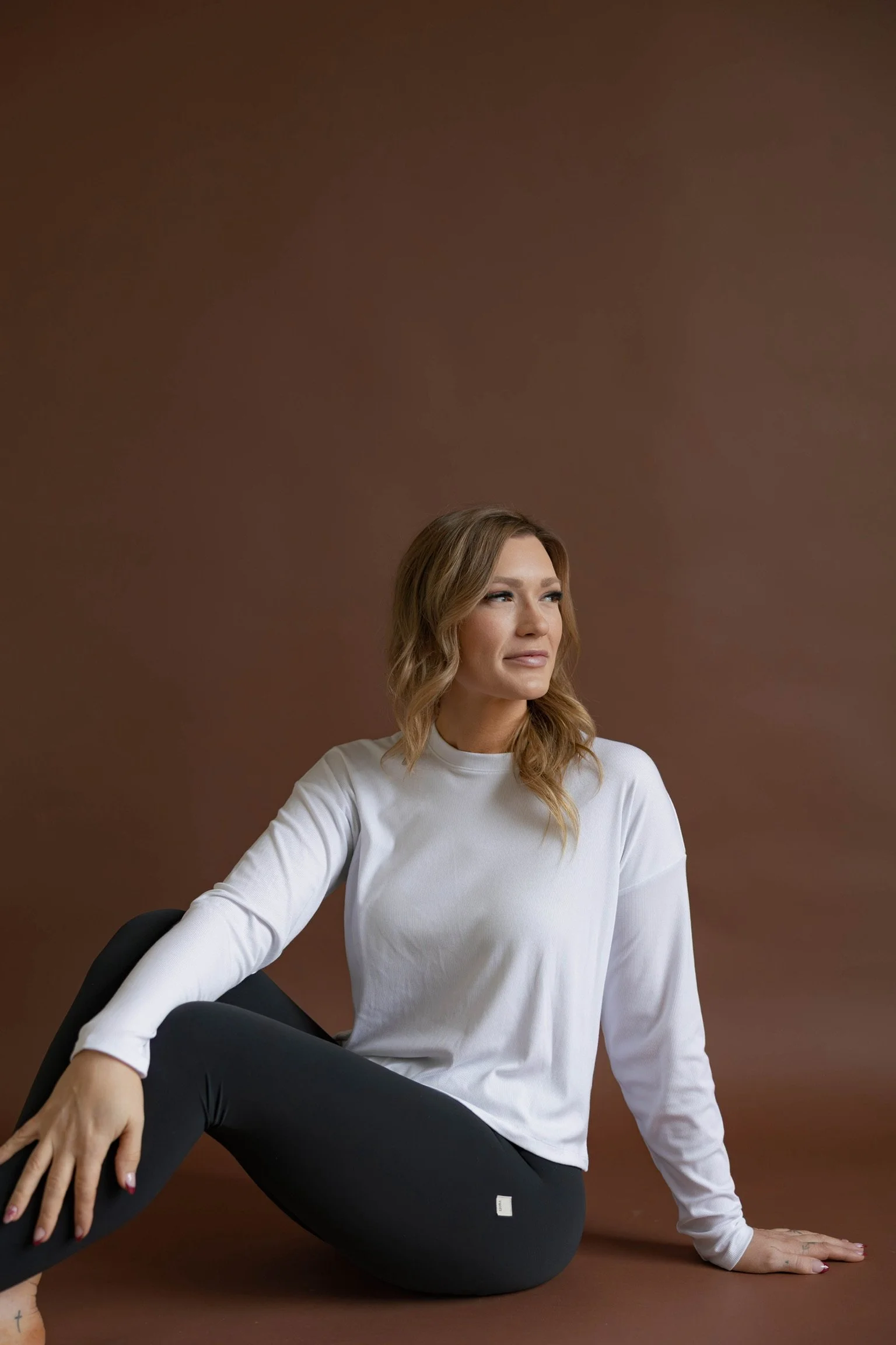 Woman with wavy light brown hair sitting against a plain brown background, wearing a white long sleeve shirt and black pants, looking to her right.