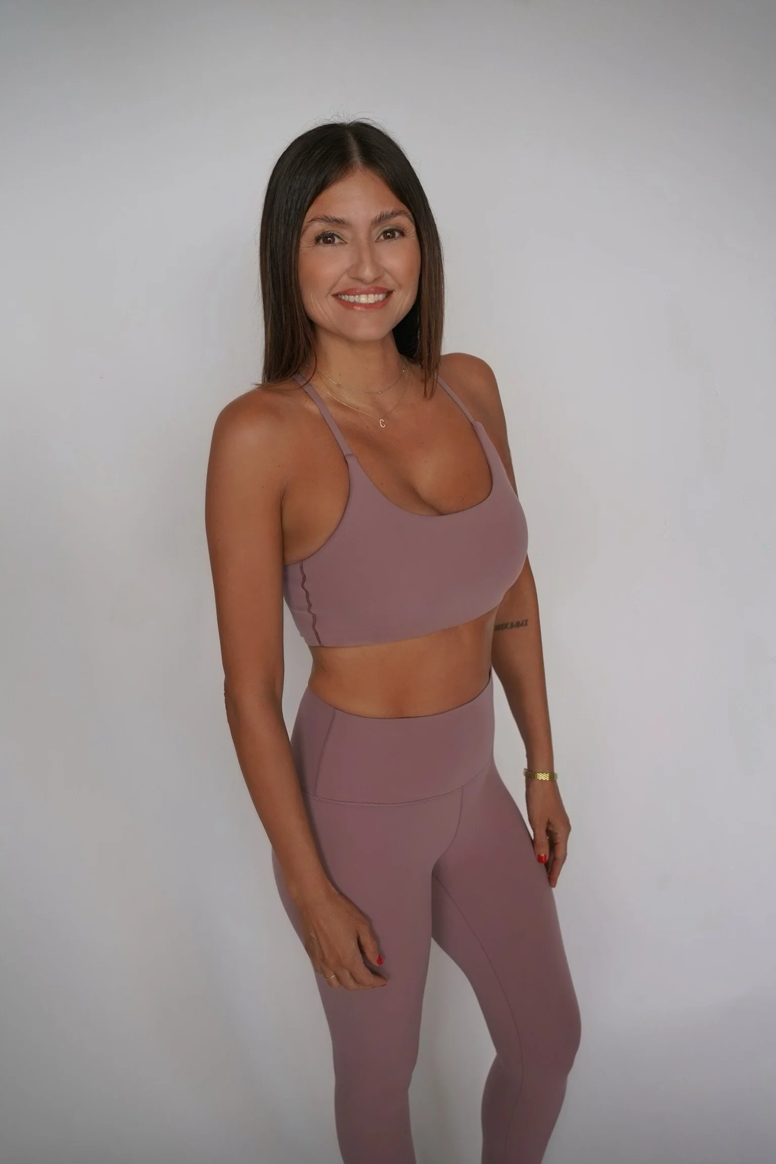 A woman with shoulder-length dark brown hair in a mauve athletic outfit, standing against a plain light gray background, smiling at the camera.