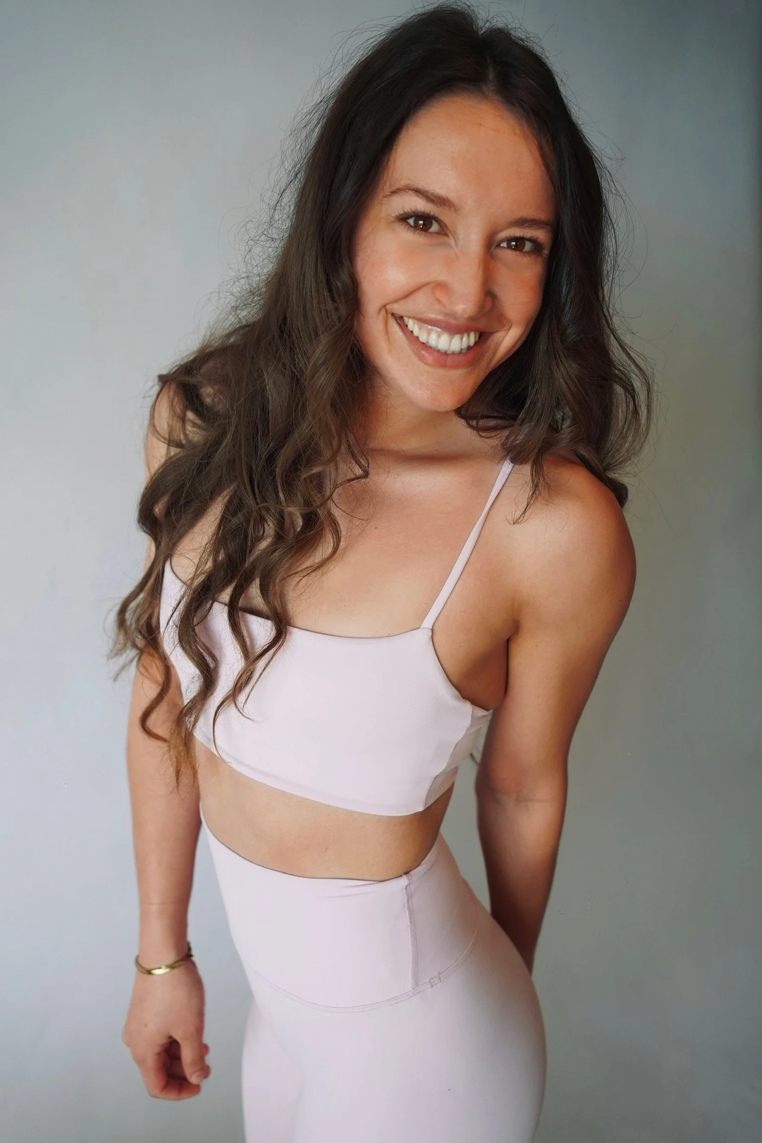 A smiling woman with long, wavy dark hair wearing a light pink crop top and matching high-waisted leggings, standing against a plain gray background.