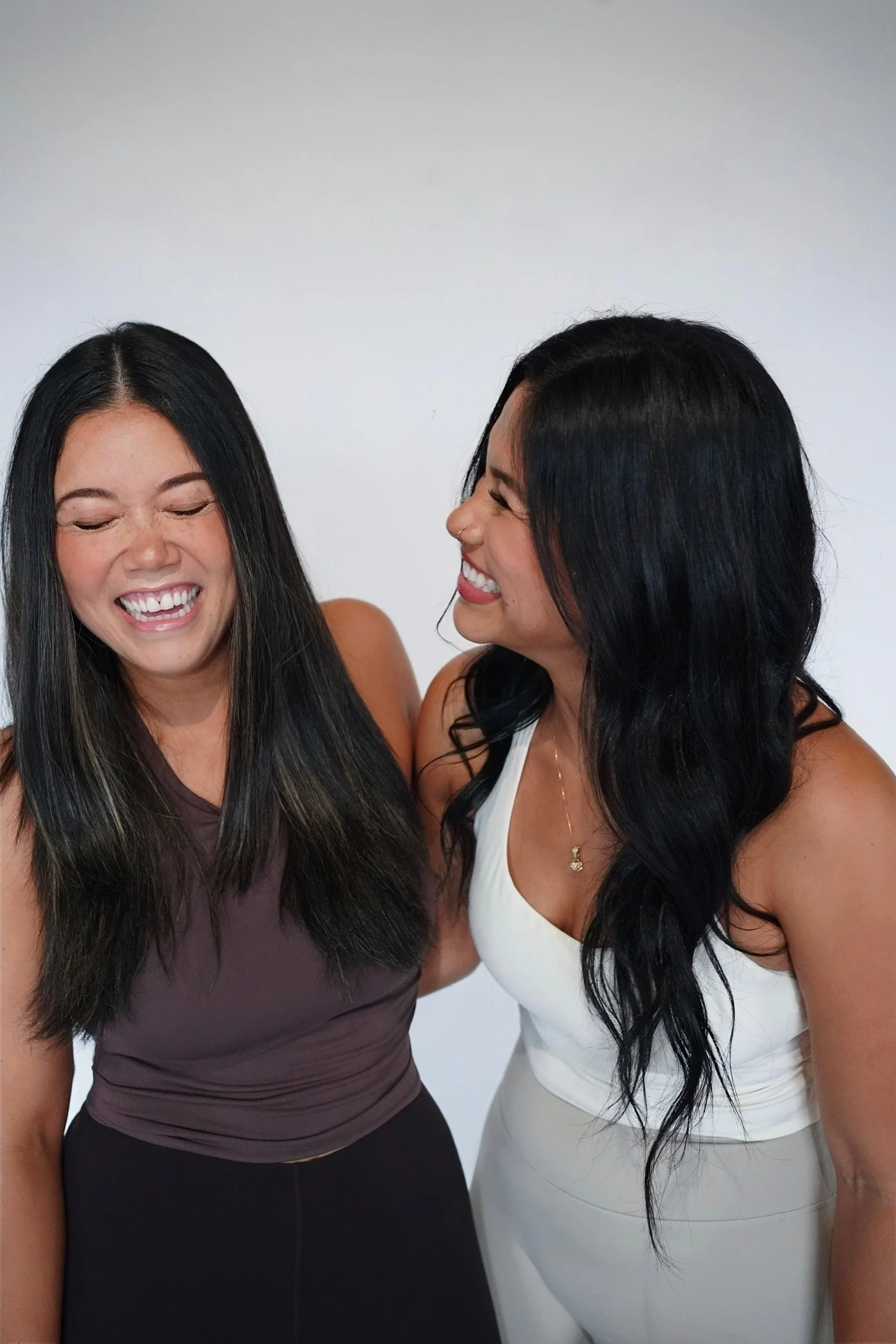 Two women laughing and smiling together against a plain background.