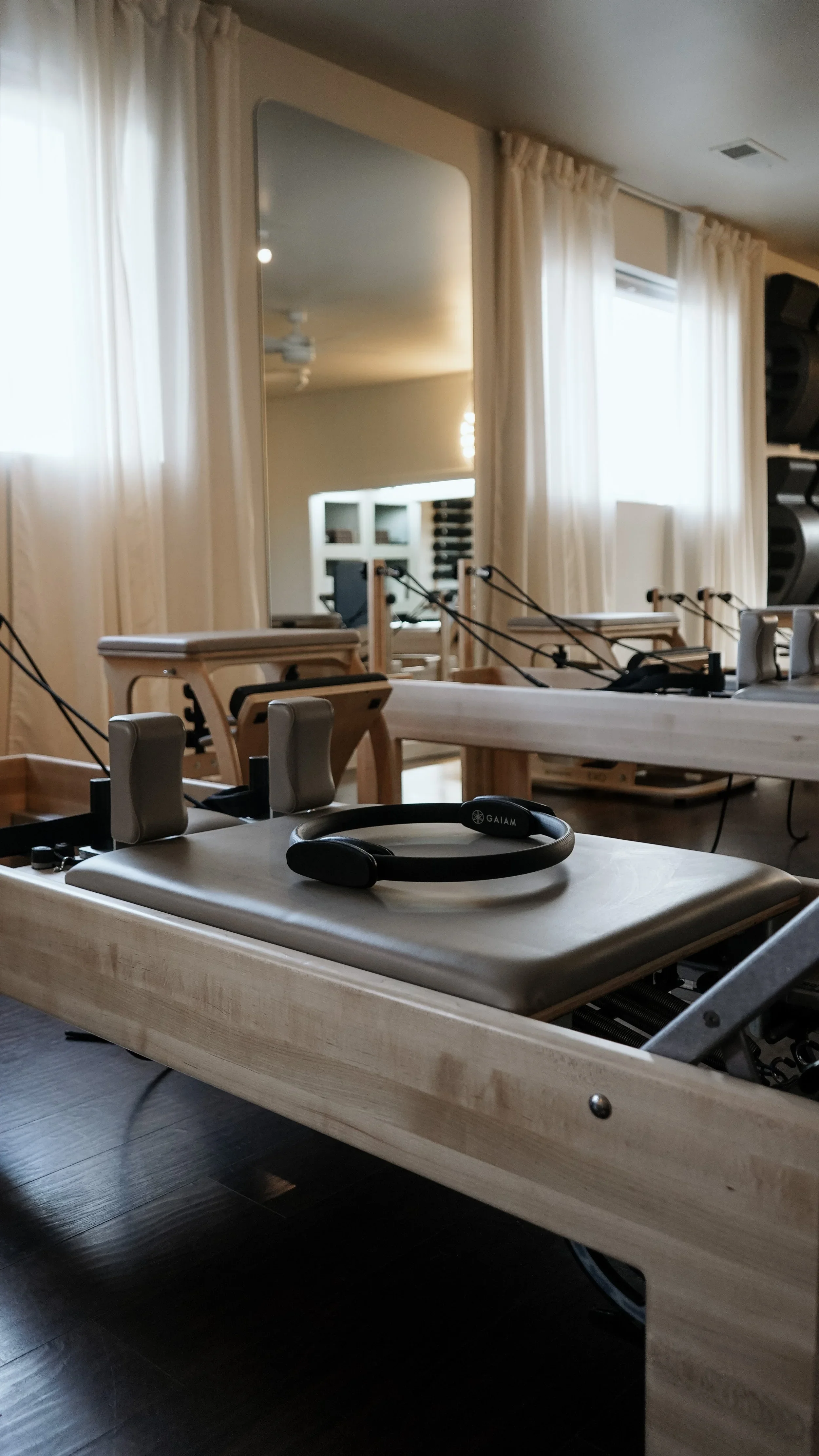Pilates reformer machine with a headband resting on it in a well-lit studio with mirrors and beige curtains.