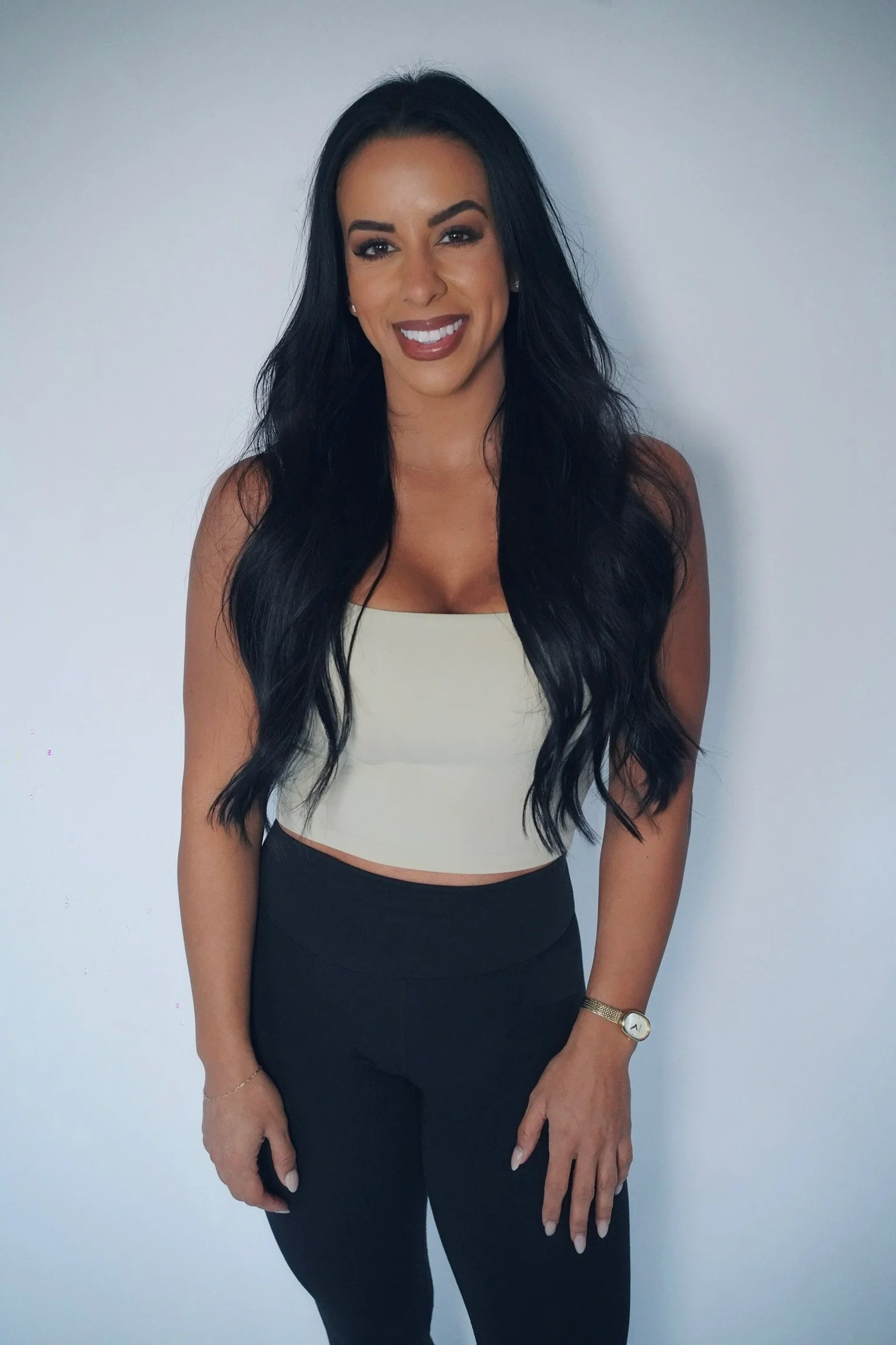 A woman with long black hair, wearing a beige crop top and black pants, standing against a plain white wall, smiling, wearing a gold watch and a bracelet.