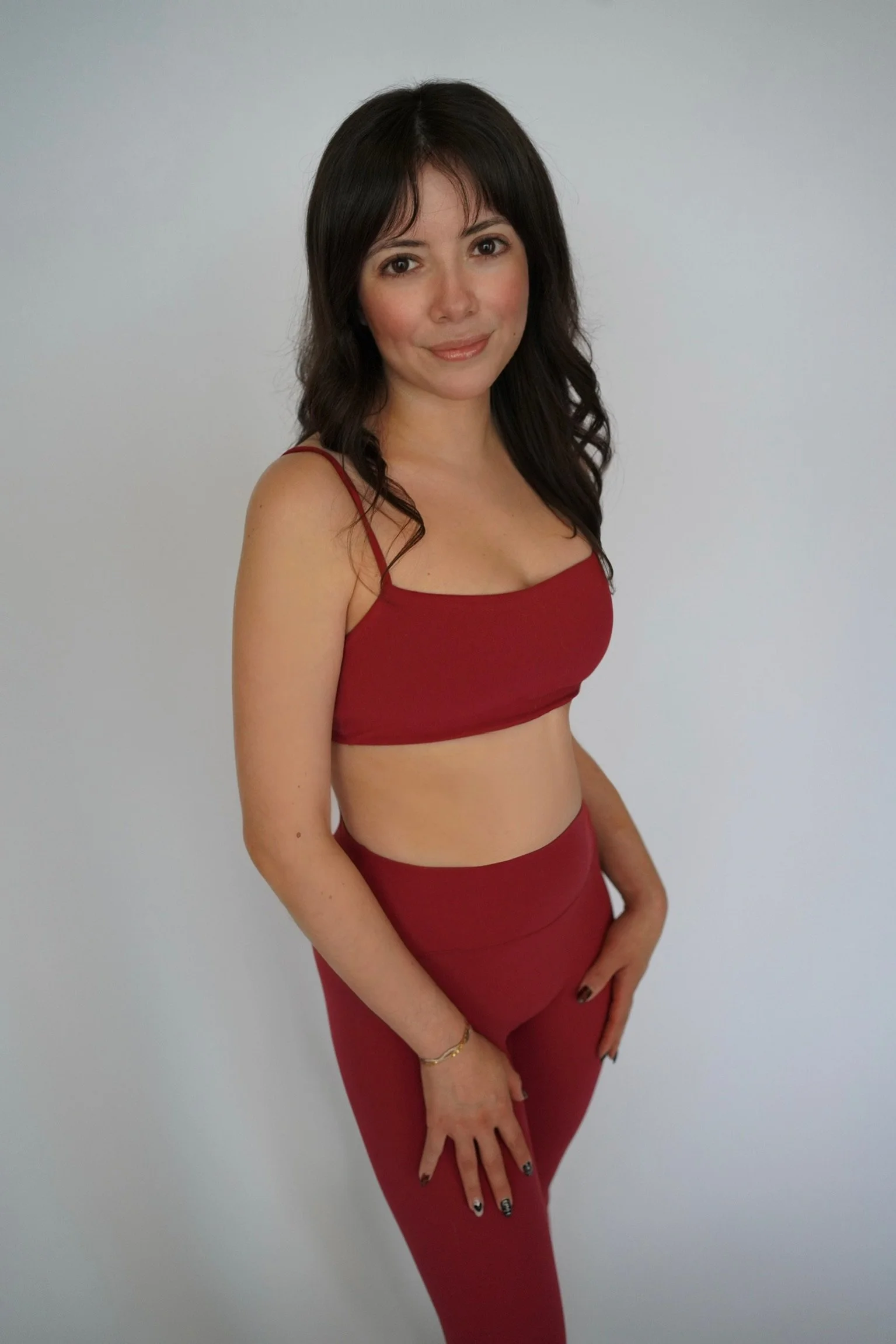 A woman wearing a red sleeveless crop top and matching high-waisted pants, with wavy dark hair, standing against a plain light-colored background.