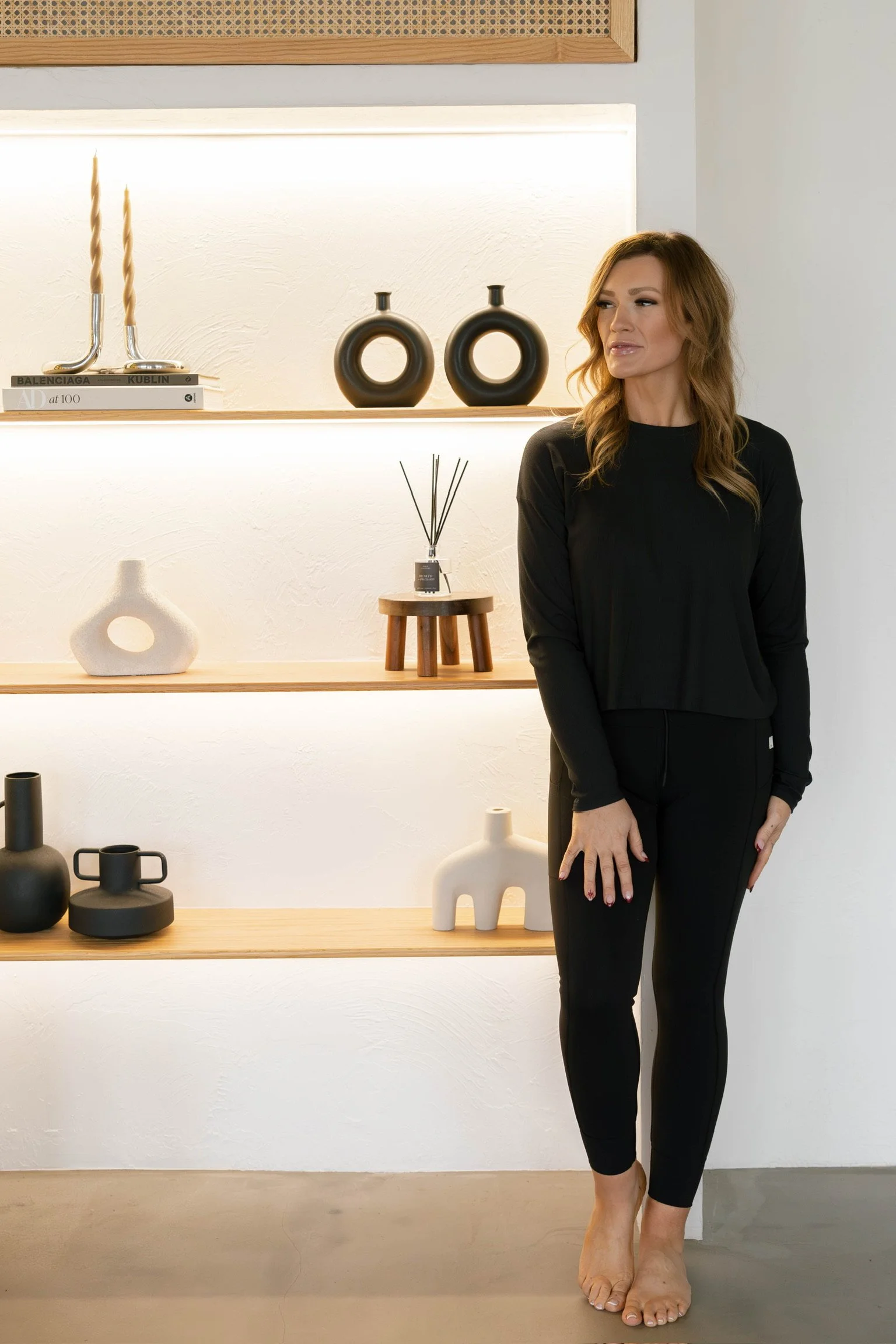 A woman standing barefoot in front of wall shelves decorated with vases, books, and reed diffuser.