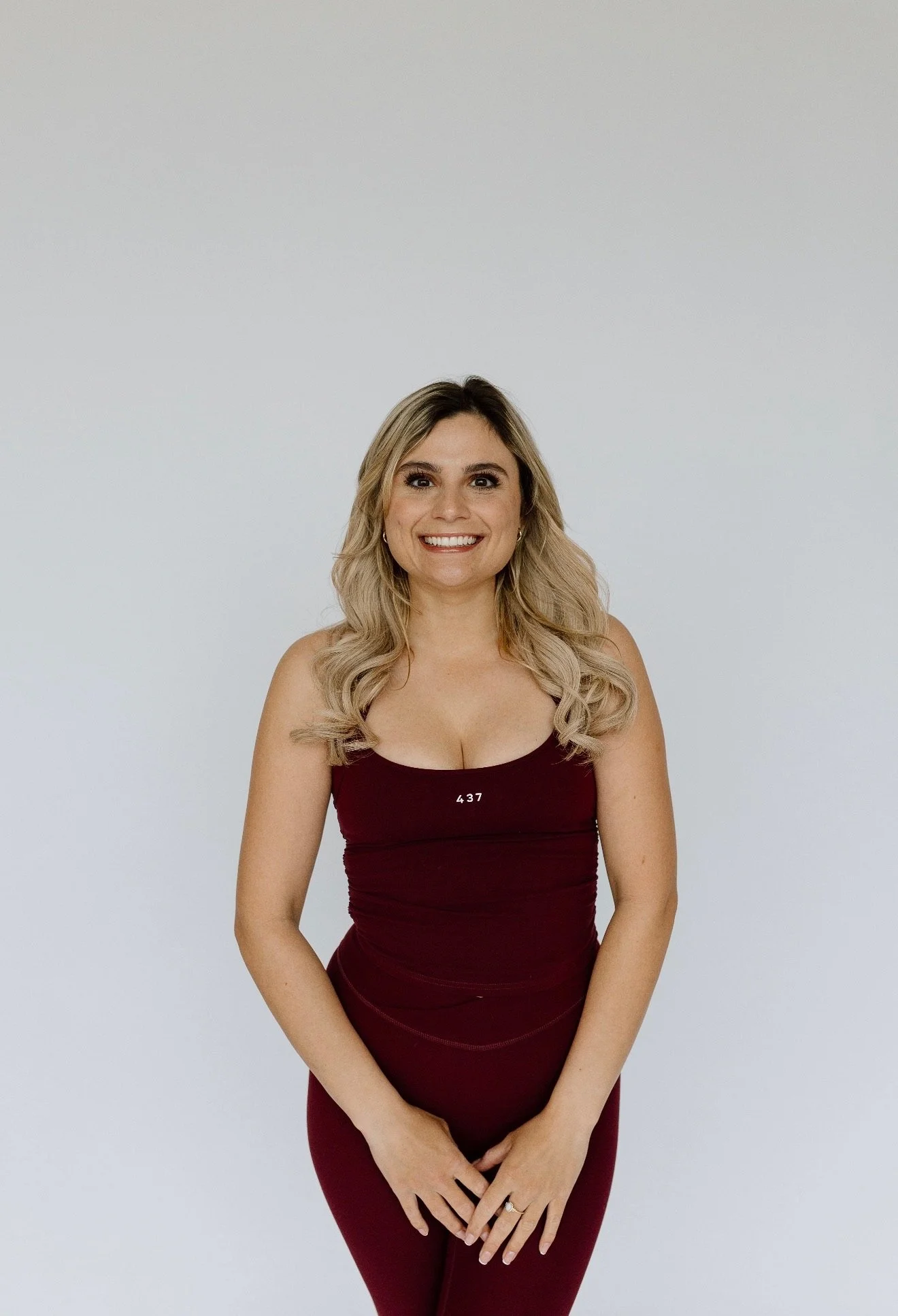 A woman with long blonde hair and a bright smile, wearing a fitted burgundy dress with the number 437 on it, standing against a plain light gray background.