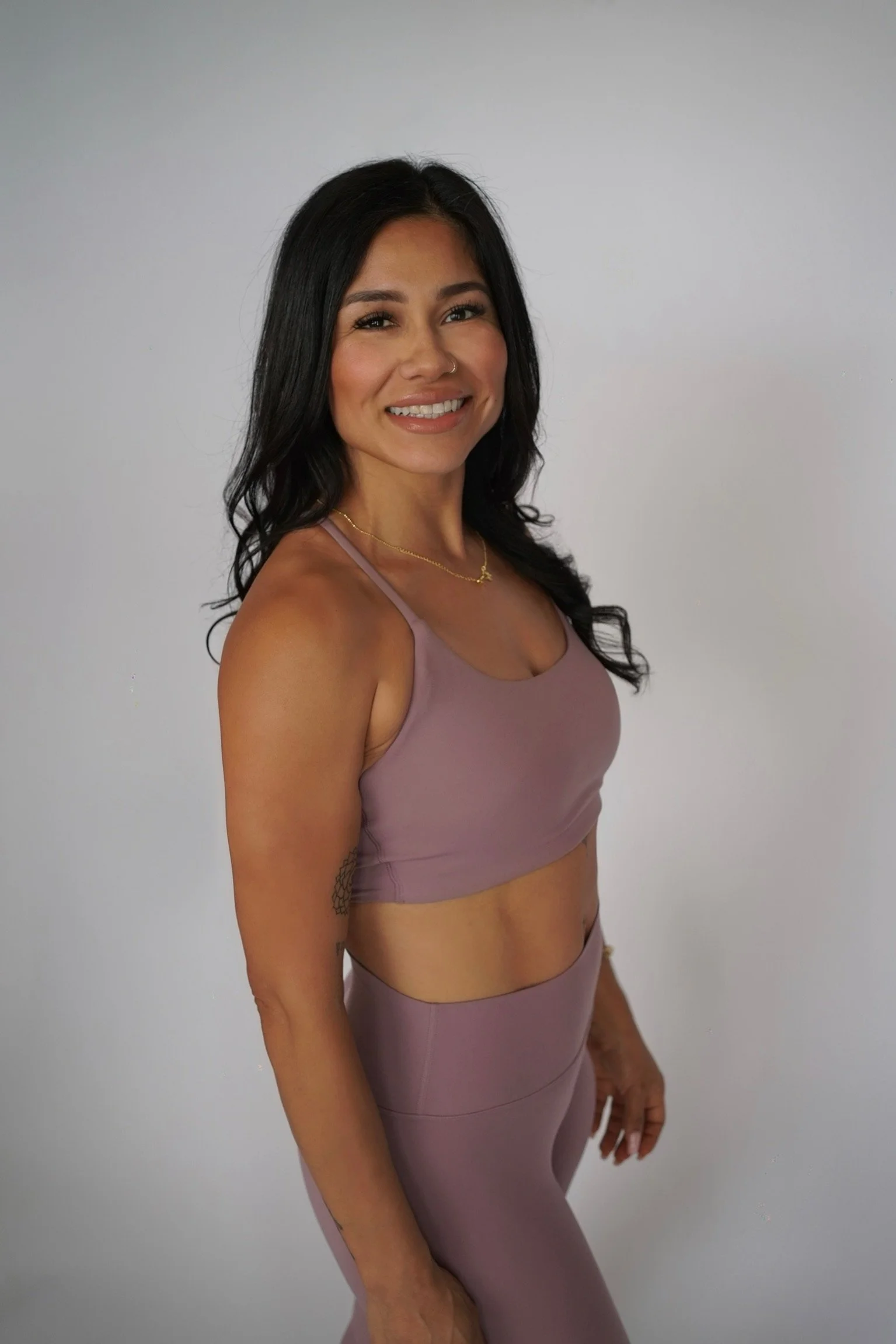 A woman with long black wavy hair smiling while wearing a mauve athletic sports bra and matching leggings, standing against a plain light gray background.