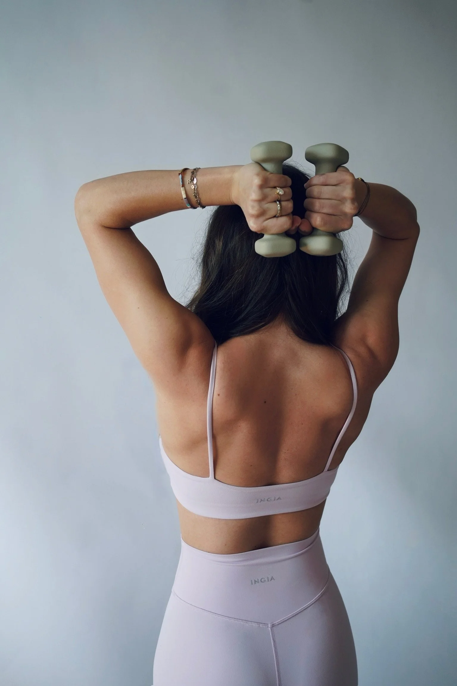 A woman in a white sports bra and high-waisted leggings holds gray dumbbells behind her head, flexing her back muscles. She is wearing jewelry on her left wrist and rings on her fingers, with her long dark hair falling forward. The background is plain and light-colored.