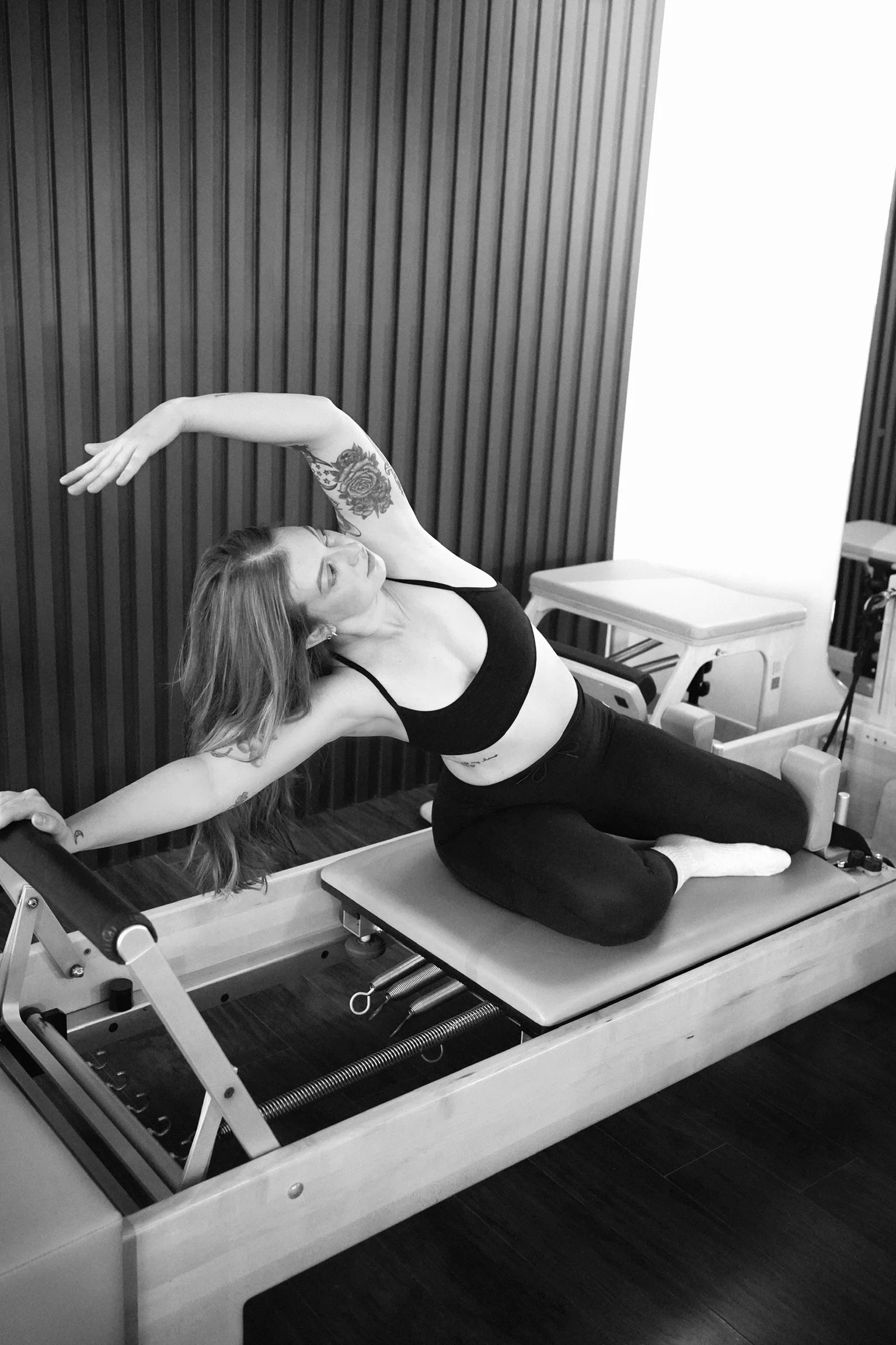 A woman practicing Pilates on a reformer machine, wearing a black sports bra and black leggings, with her hair down and a tattoo on her left arm, in a room with wood-paneled wall.