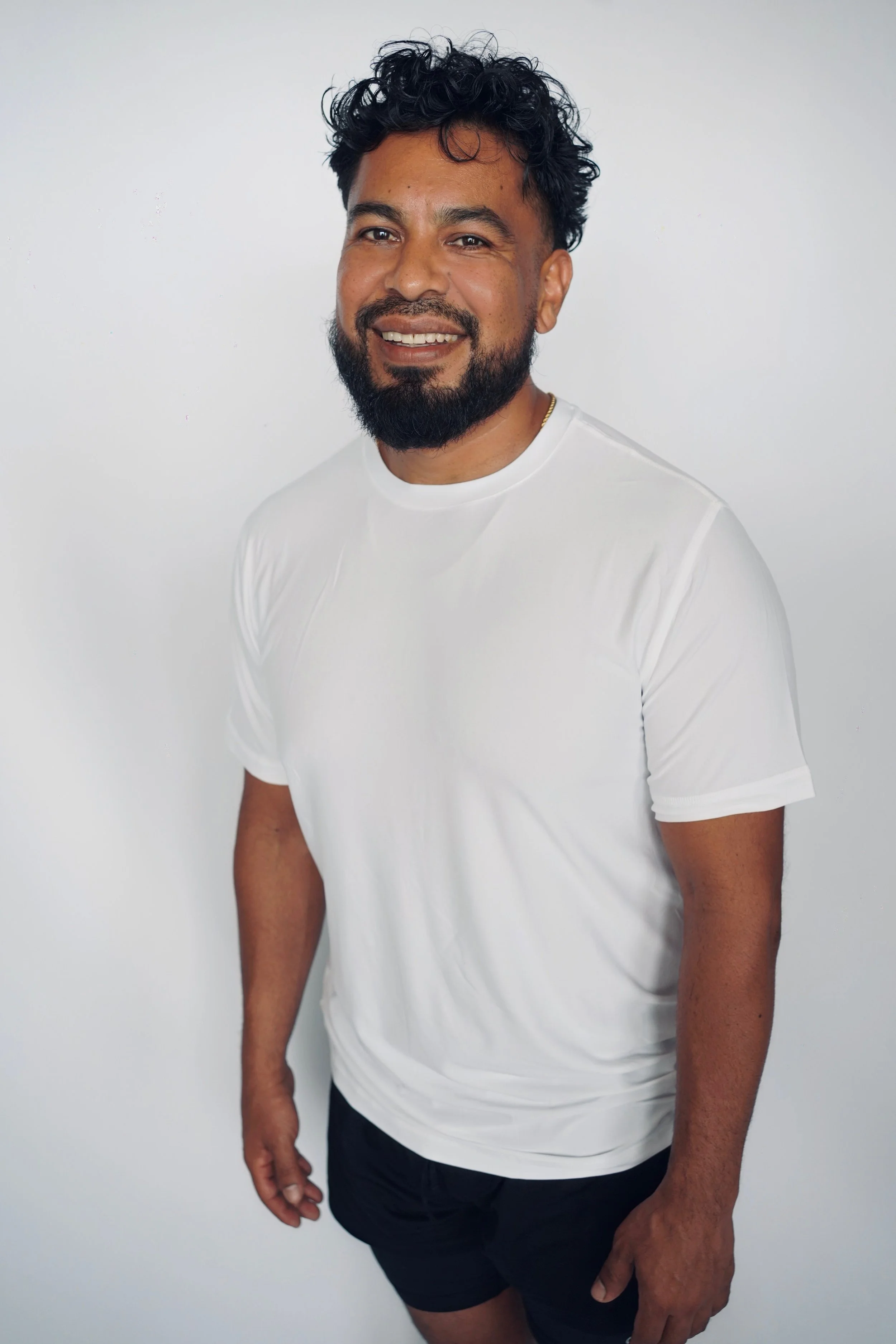 A man with curly black hair and a beard, wearing a white t-shirt and dark shorts, standing against a plain white background and smiling at the camera.