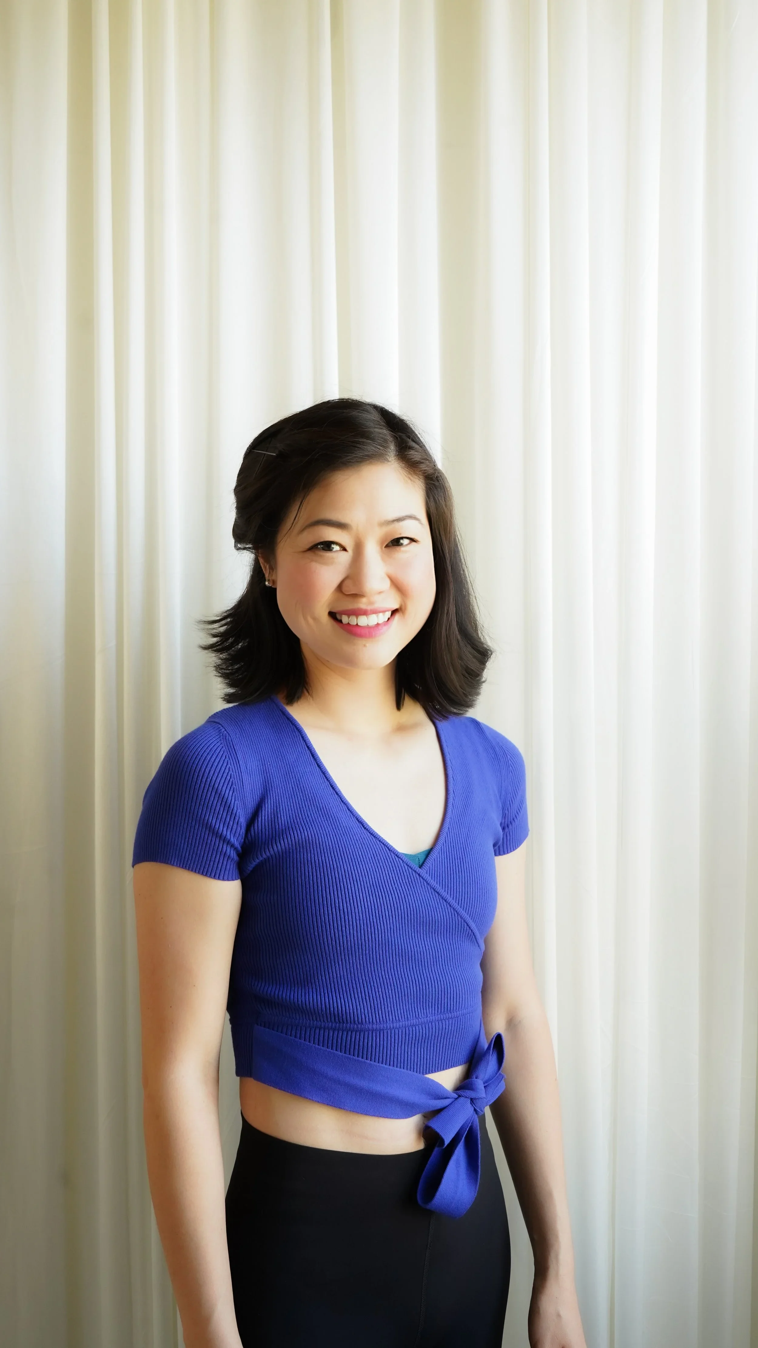 A young woman with shoulder-length dark hair, smiling, wearing a bright blue, short-sleeved, wrap-knit crop top with a bow tied at the waist, and black pants, standing in front of off-white curtains.