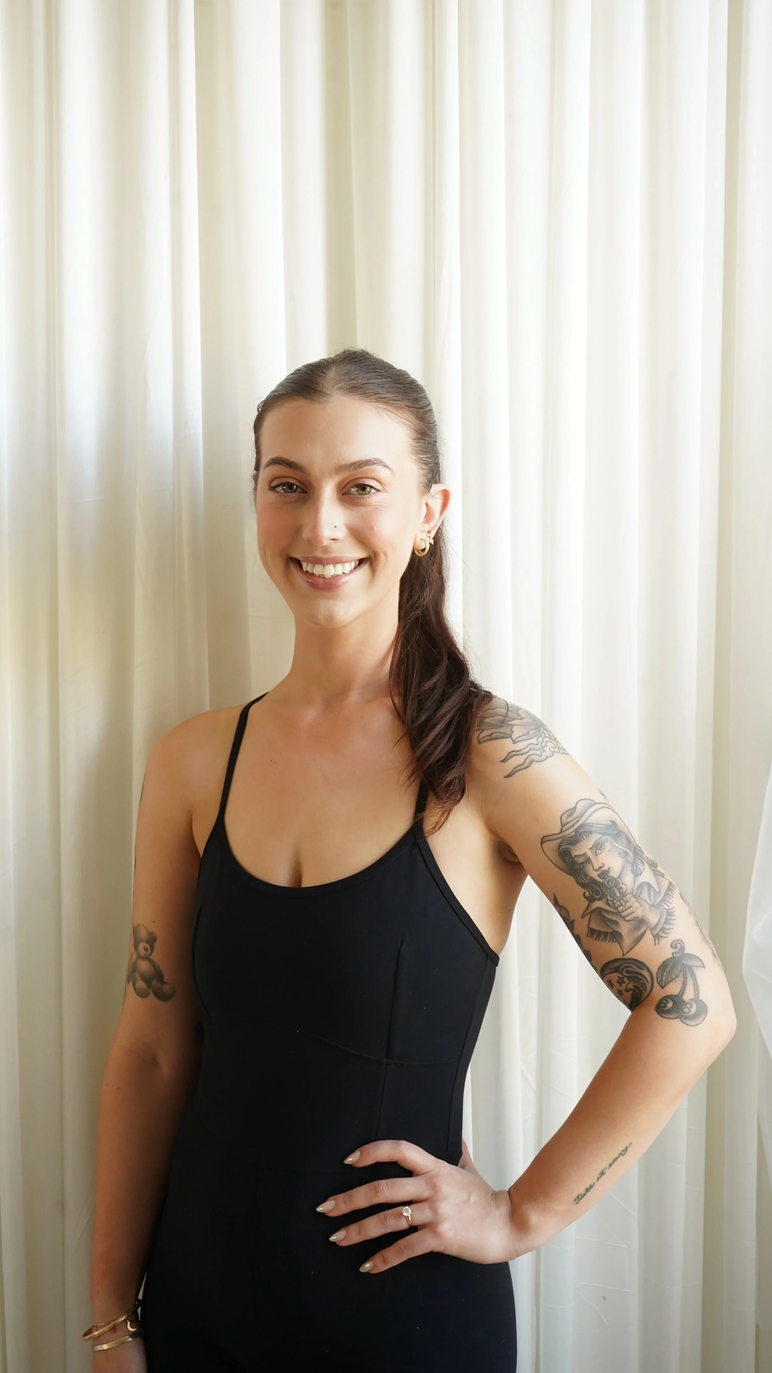 A smiling woman in a black tank top standing in front of cream-colored curtains.