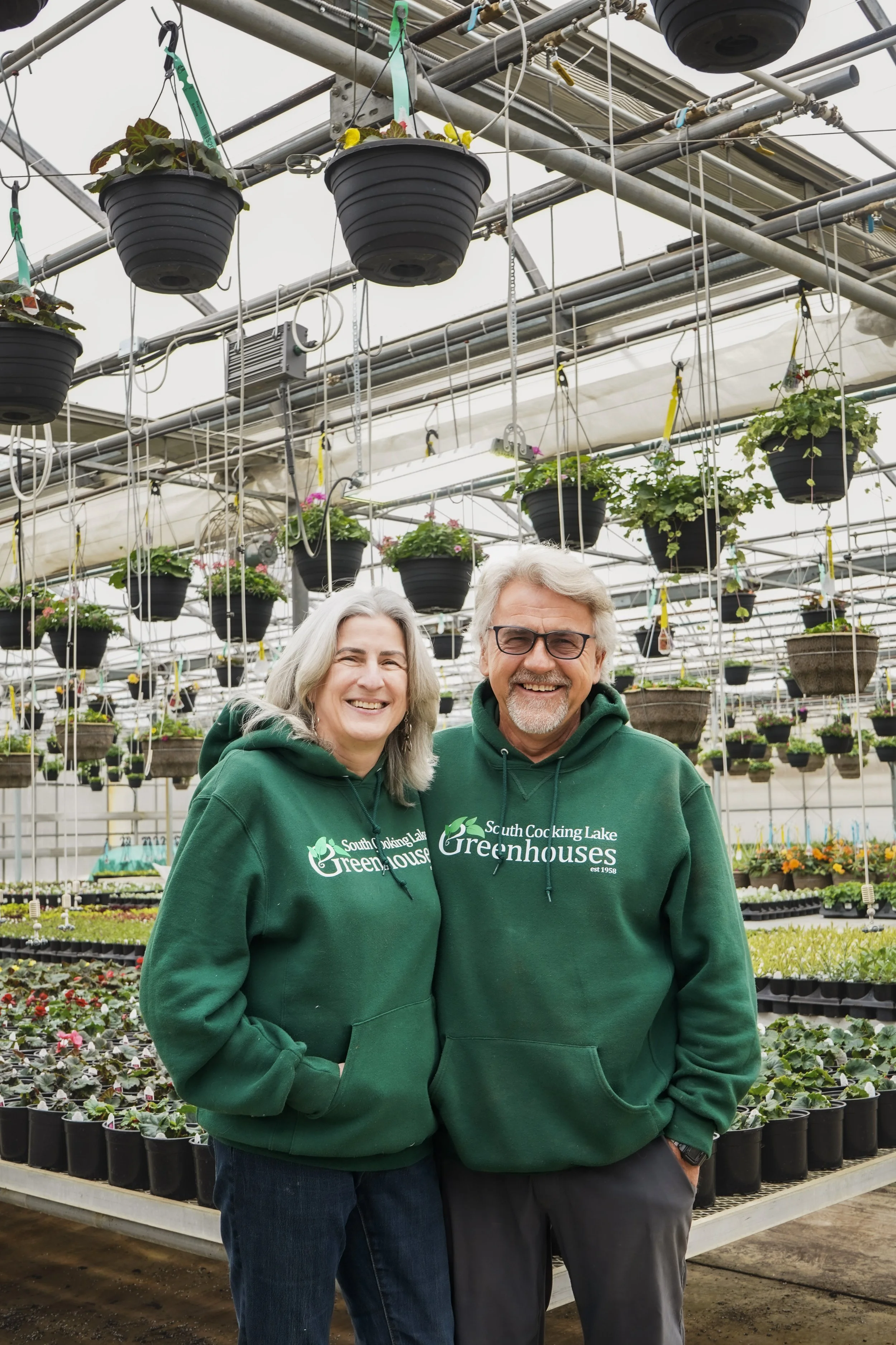 A smiling woman and man wearing green hoodies that say 'South Cooking Lake Greenhouses' are standing inside a greenhouse filled with hanging pots and potted plants.