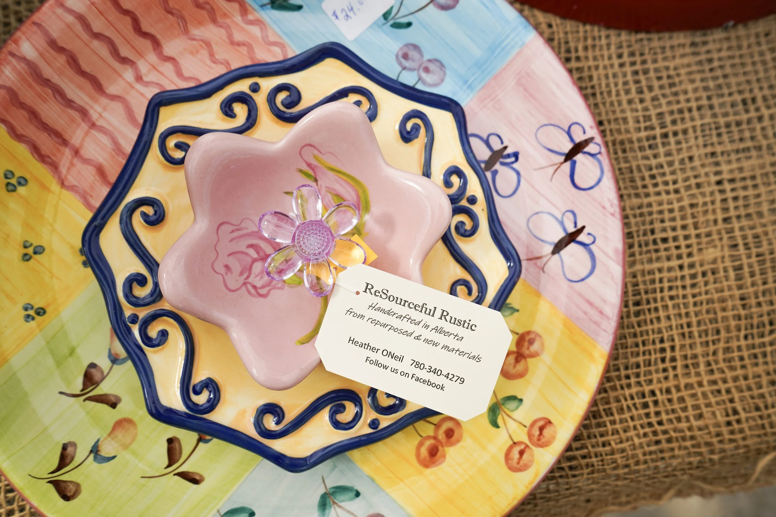 Colorful ceramic plate with intricate blue swirling patterns, holding a pink handcrafted soap shaped like a flower with a decorative clear flower on top. The plate and soap sit on a vibrant, multi-colored tablecloth with floral and abstract designs, with a business card partially visible underneath.