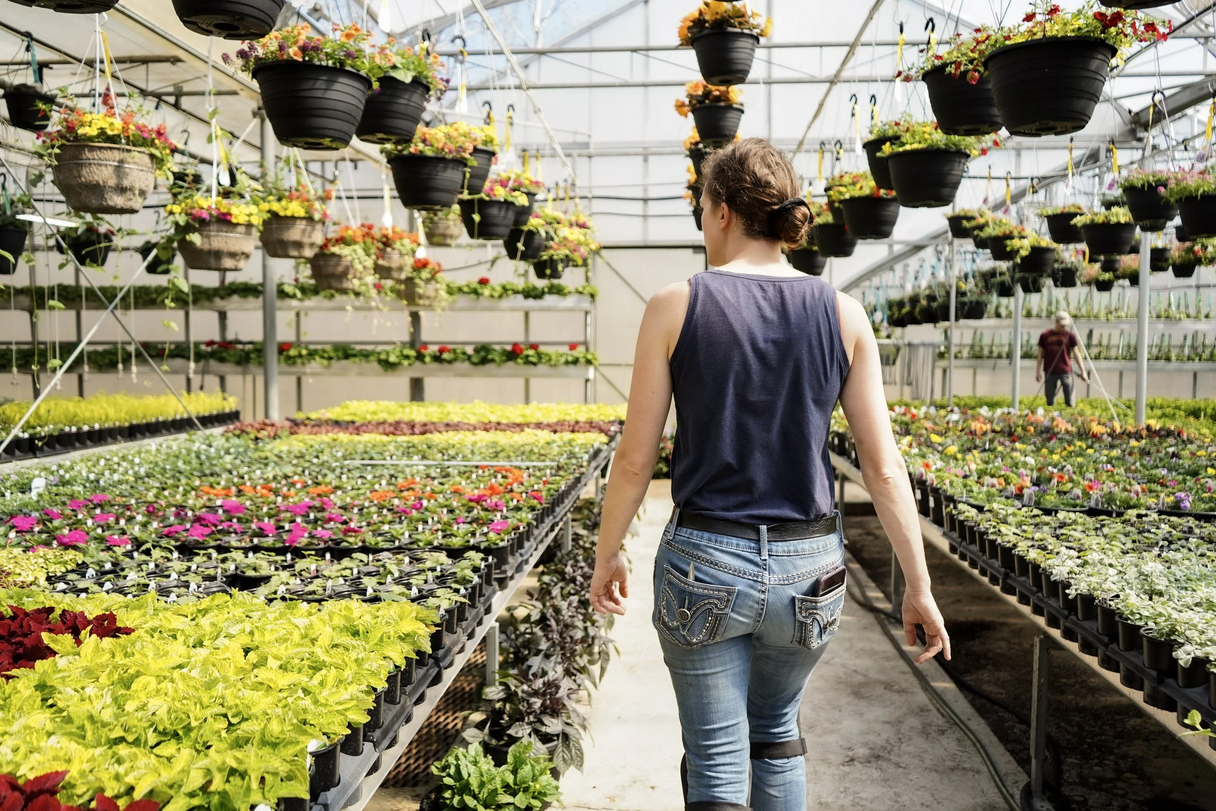 A woman in a blue sleeveless shirt and jeans walking in a greenhouse or plant nursery surrounded by shelves of colorful flowering plants and hanging flower pots.