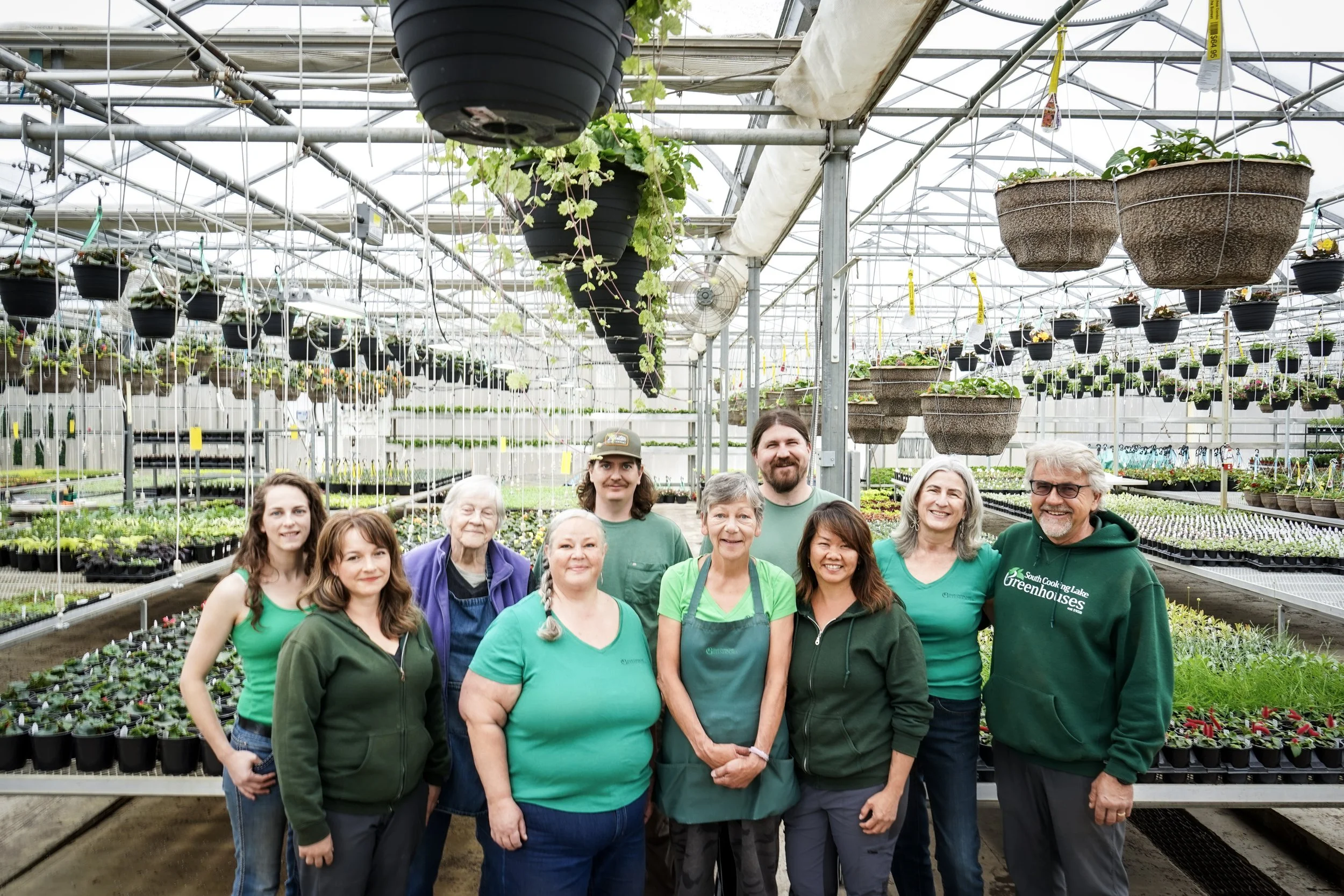 Group of diverse people in green and blue shirts inside a greenhouse, surrounded by hanging pots and plant rows.