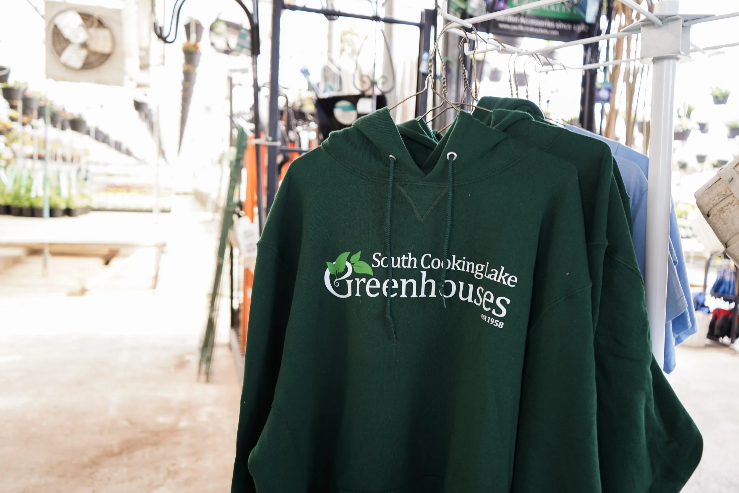 Green hoodie with white text that reads 'South Cooking Lake Greenhouses est 1958' hanging on a clothing rack in a greenhouse or garden center.