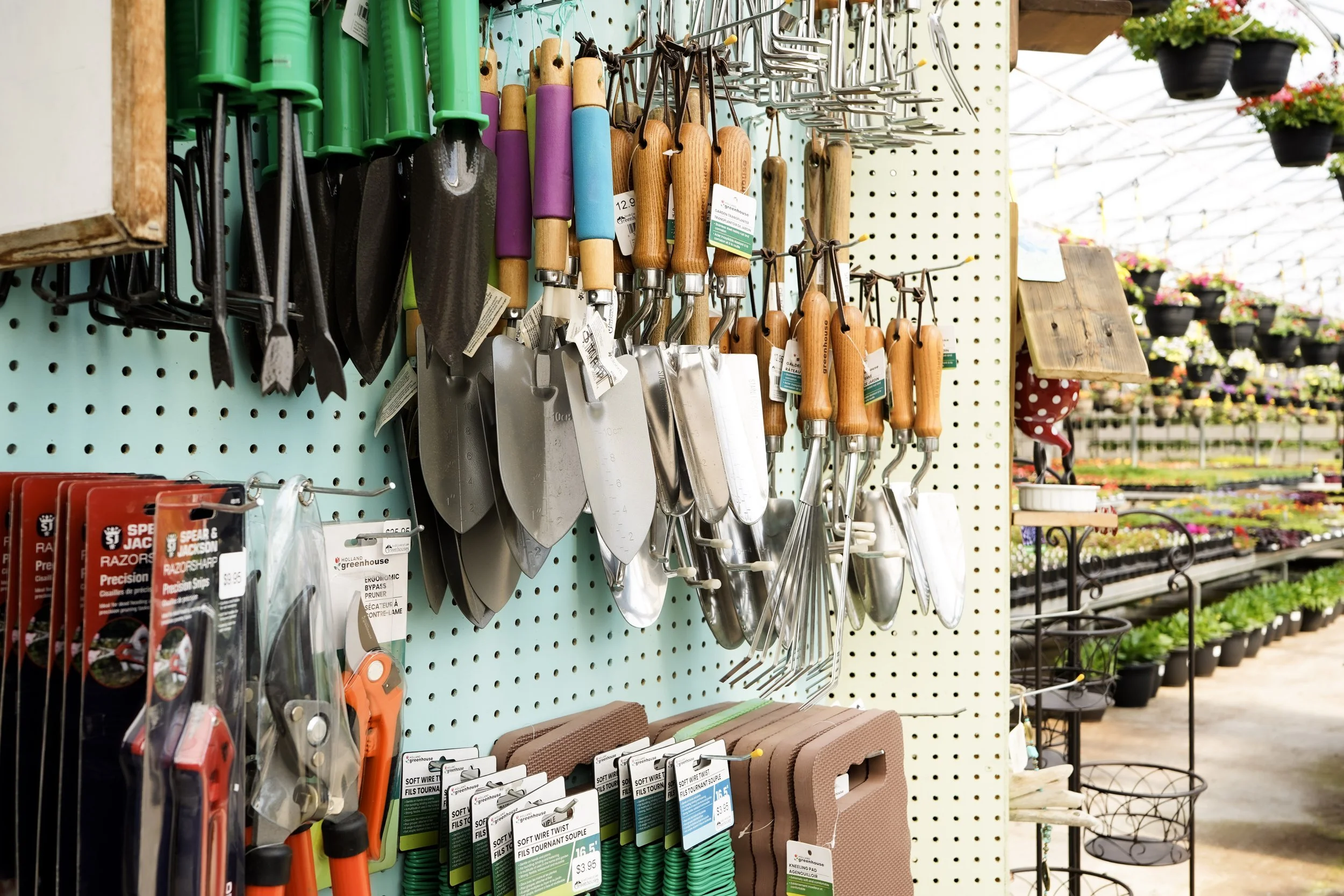 A display of gardening tools and supplies in a garden center, including hand trowels, transplanters, and garden kneeling pads, with shelves of flowers and plants in the background.